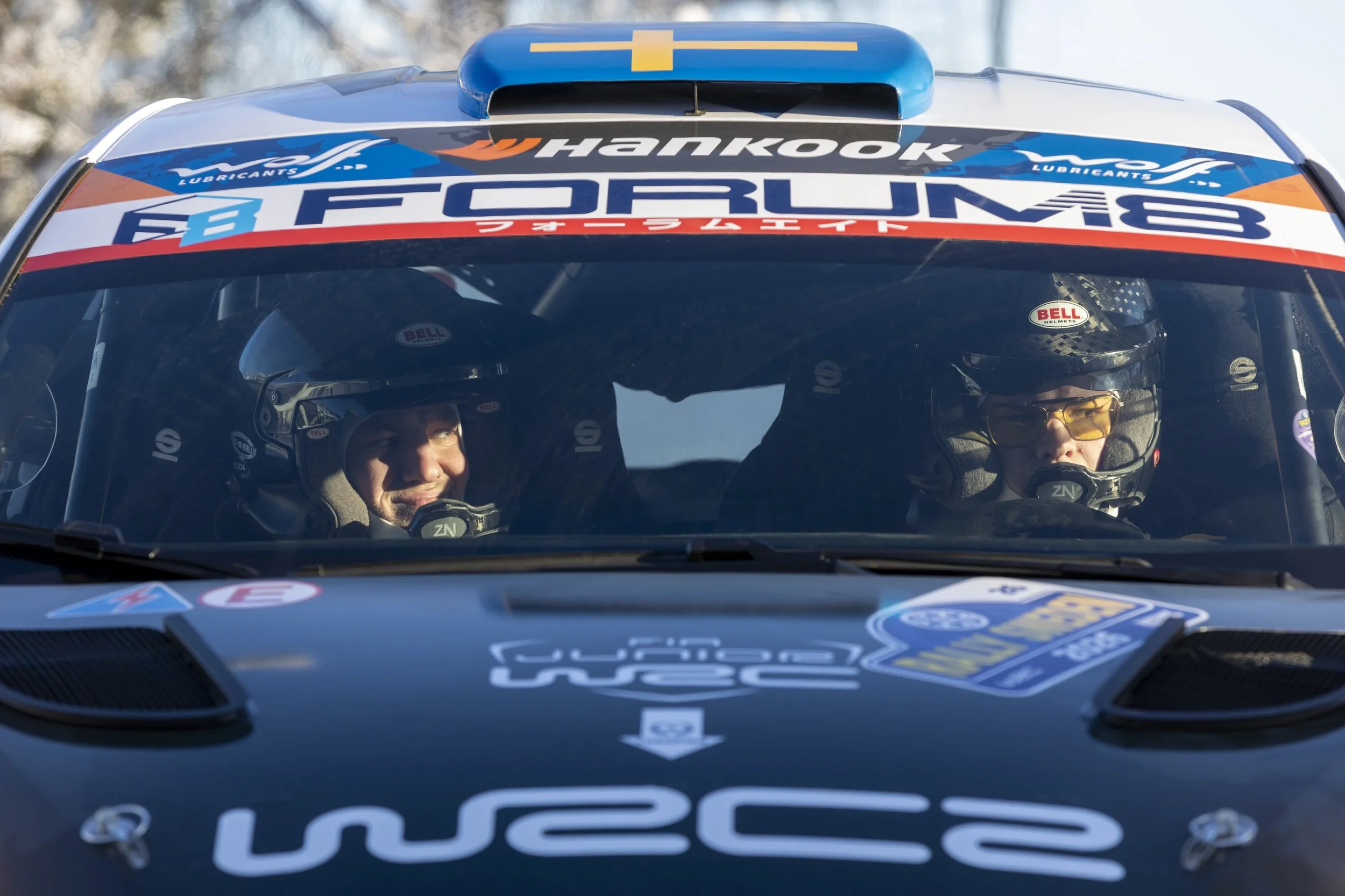 Close-up of a rally car's windshield showing two drivers wearing helmets and racing suits inside, adorned with various sponsor logos, including Hankook and Ford M.