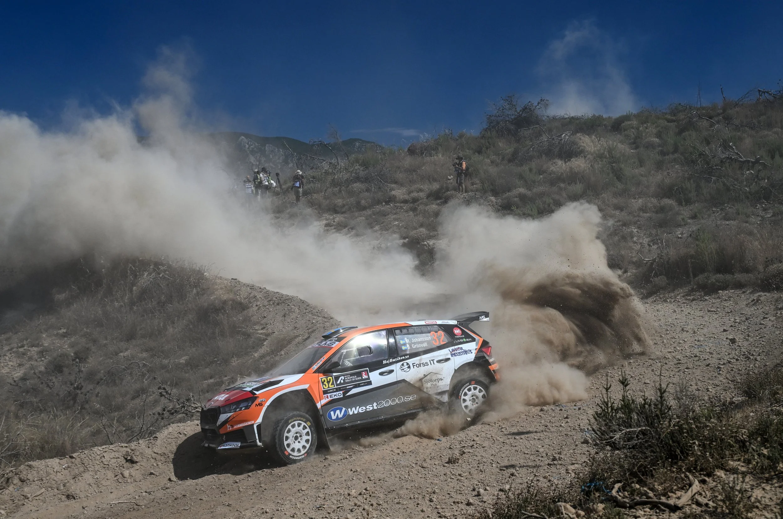A rally car with the number 32 speeds through a dusty off-road trail, kicking up a large cloud of dust behind it, with a mountain landscape and blue sky in the background.