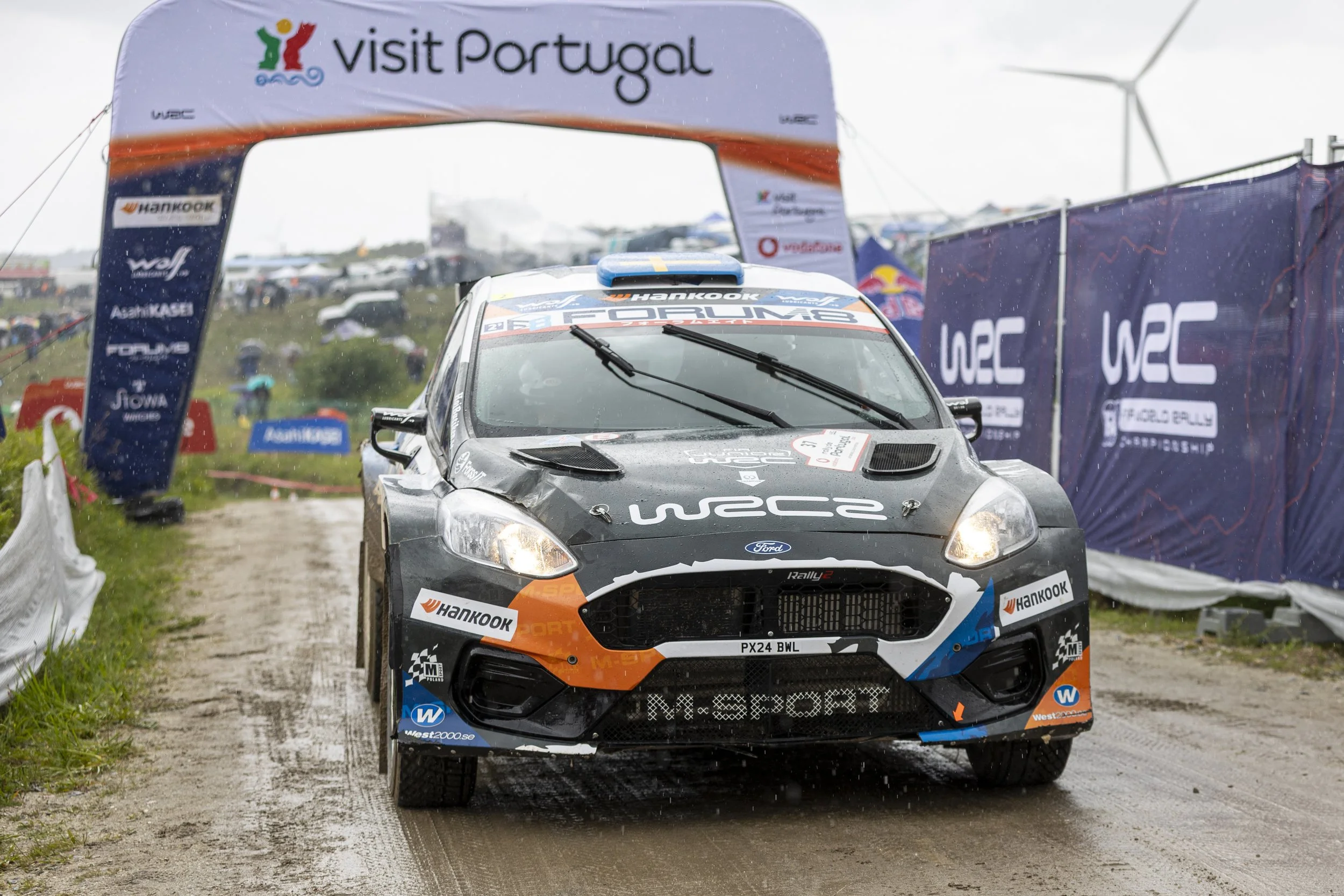 A rally car crossing the finish line under a 'Visit Portugal' banner on a rainy day, with spectators and wind turbines in the background.