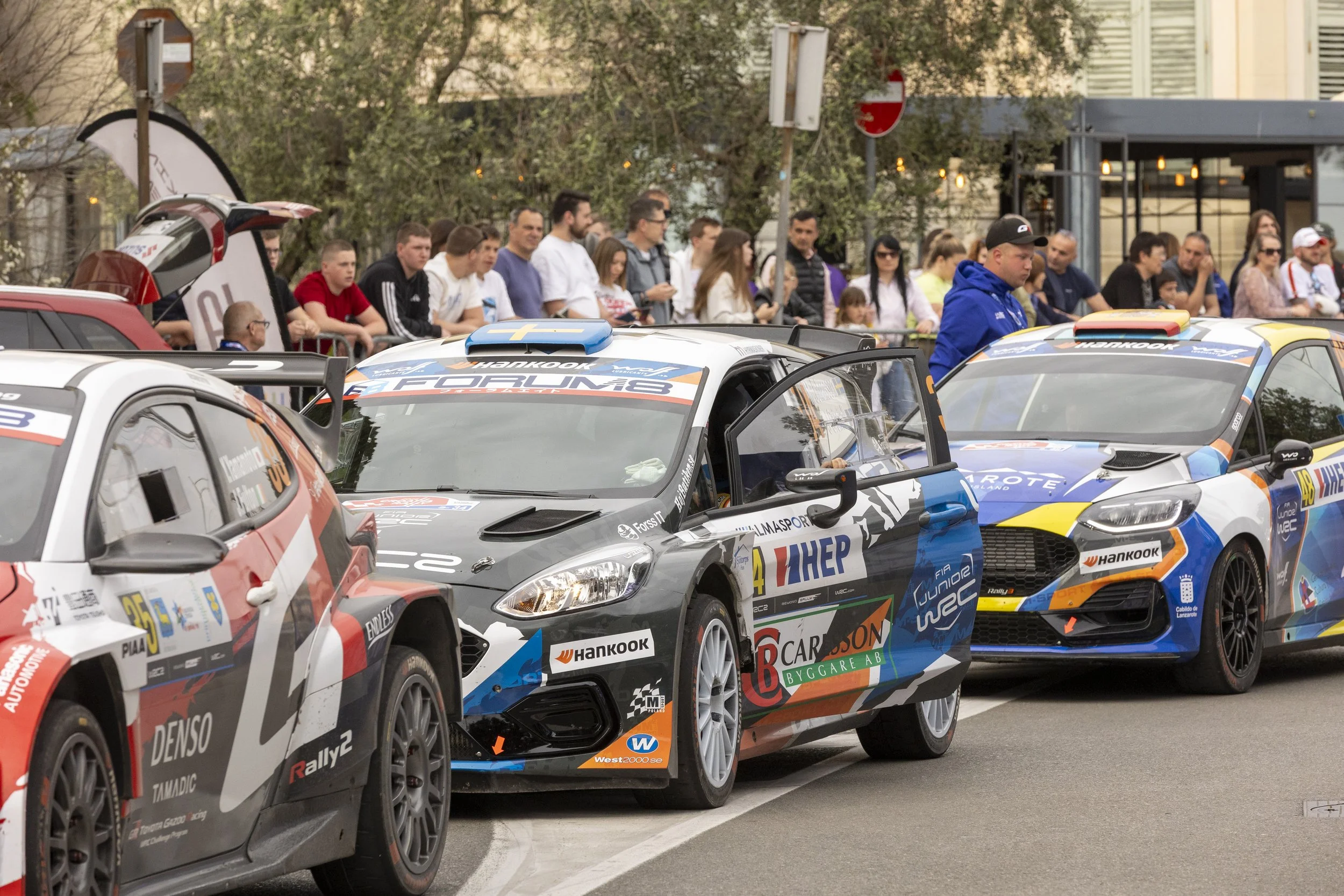 Multiple rally cars parked in a row, with a crowd of spectators behind a barrier watching the event. The scene appears to be at a rally racing event.