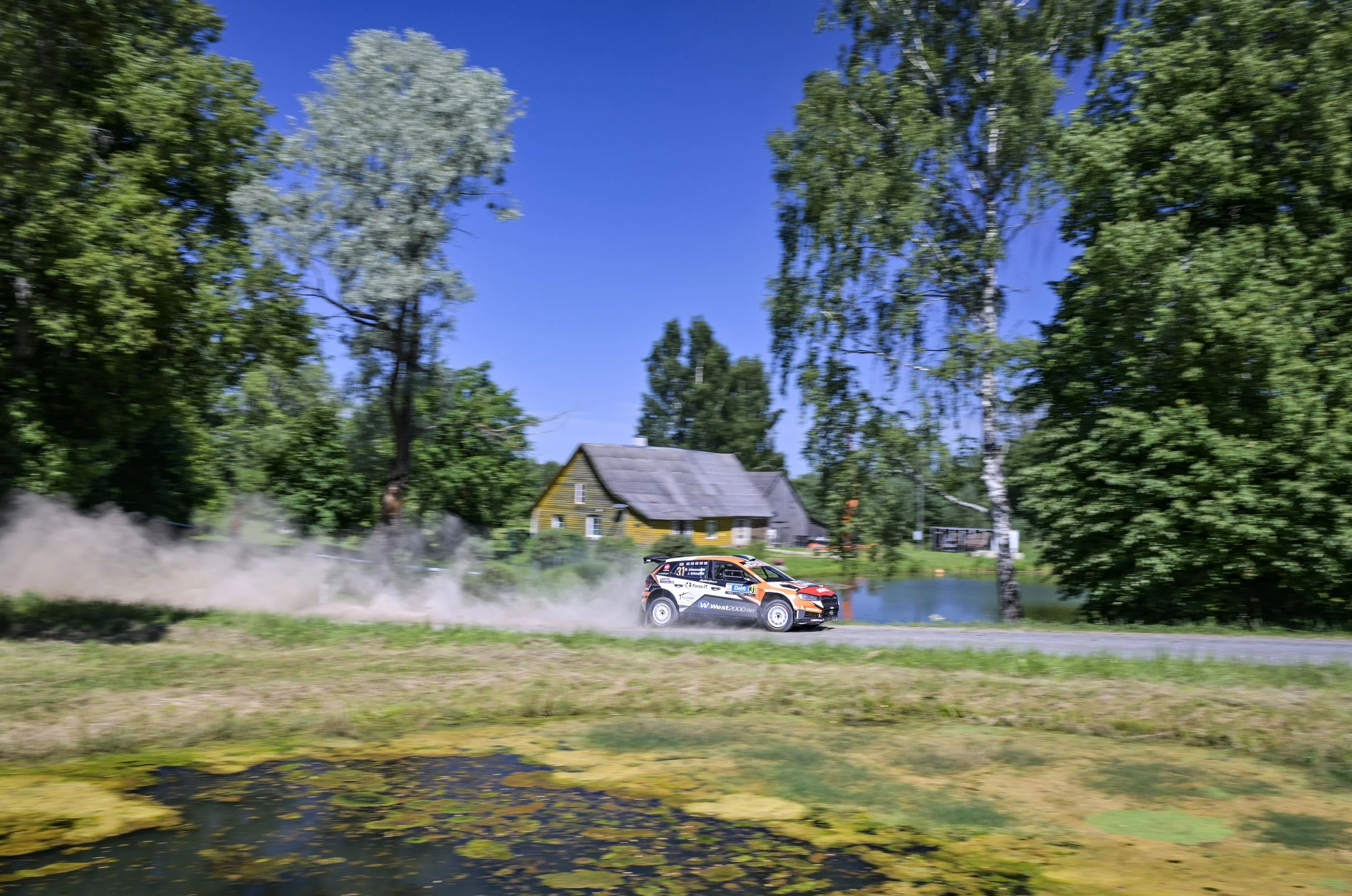 A rally car speeding along a rural road, kicking up dust behind it, with green trees, a house, and a pond in the background under a blue sky.