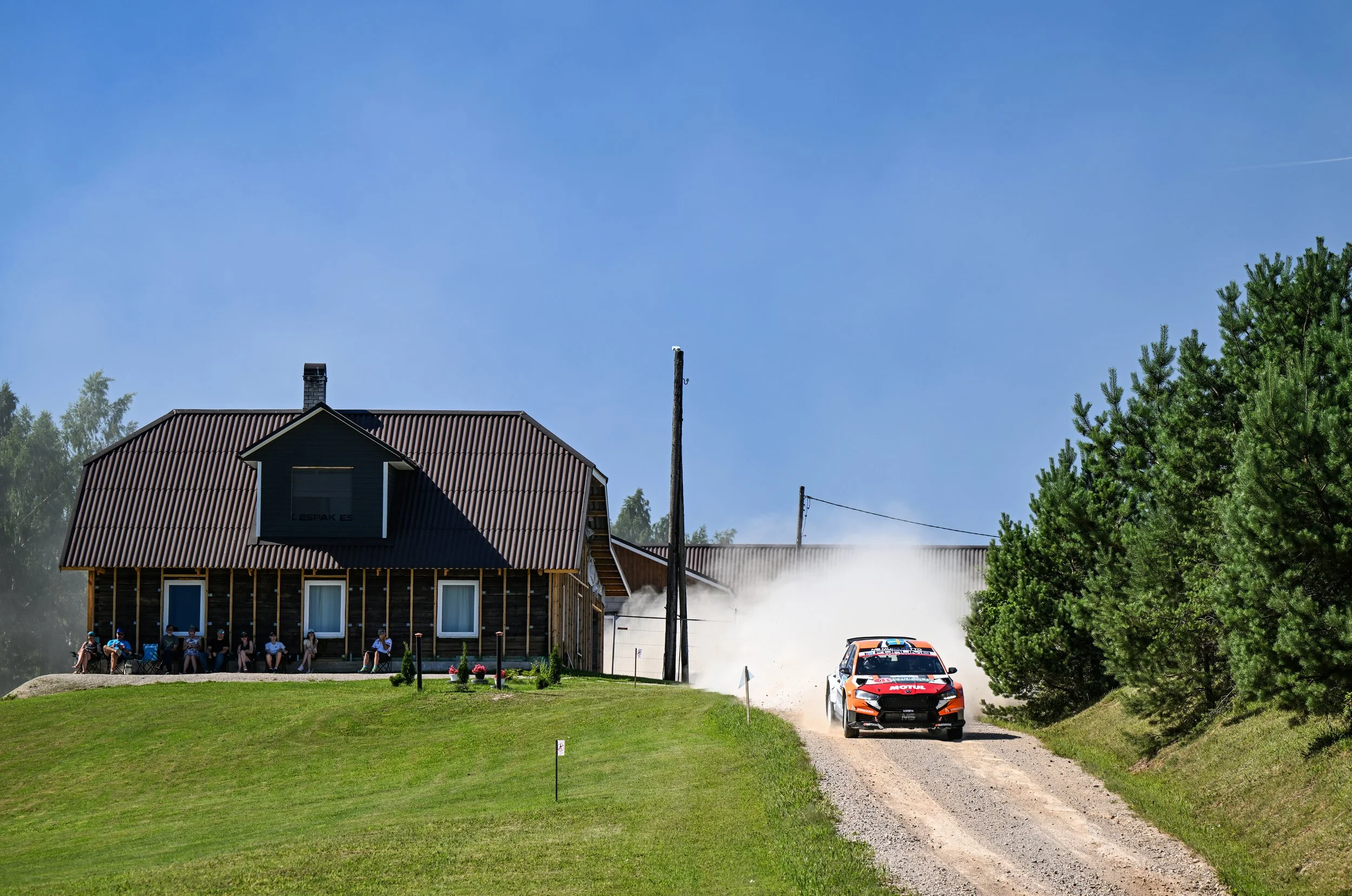 A rally car racing on a gravel driveway in front of a house, with a group of people sitting on the porch. Smoke is visible behind the car, with trees and a blue sky in the background.