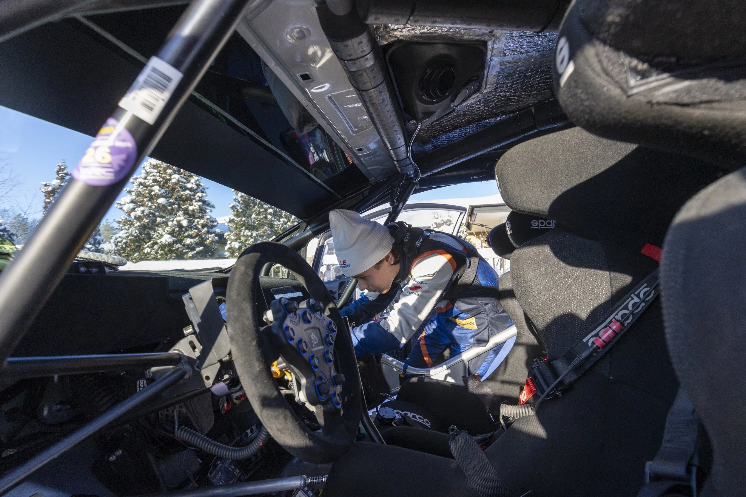 A race car driver in racing gear, including a white hat and racing suit, working inside the cockpit of a rally car. The car's interior with equipment and a roll cage is visible, with trees and snow outside the open door.