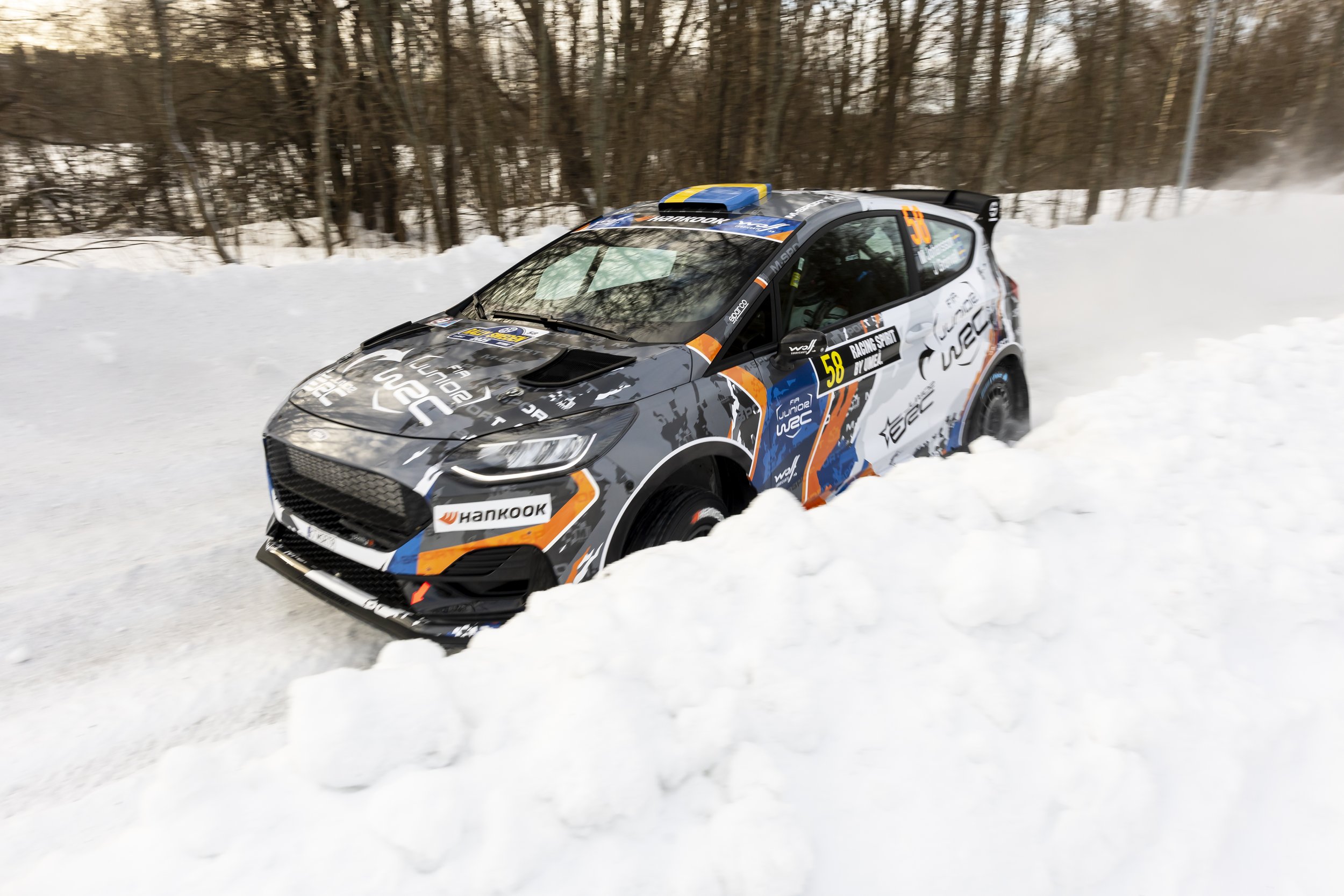 A rally car speeds through a snowy landscape with trees in the background.
