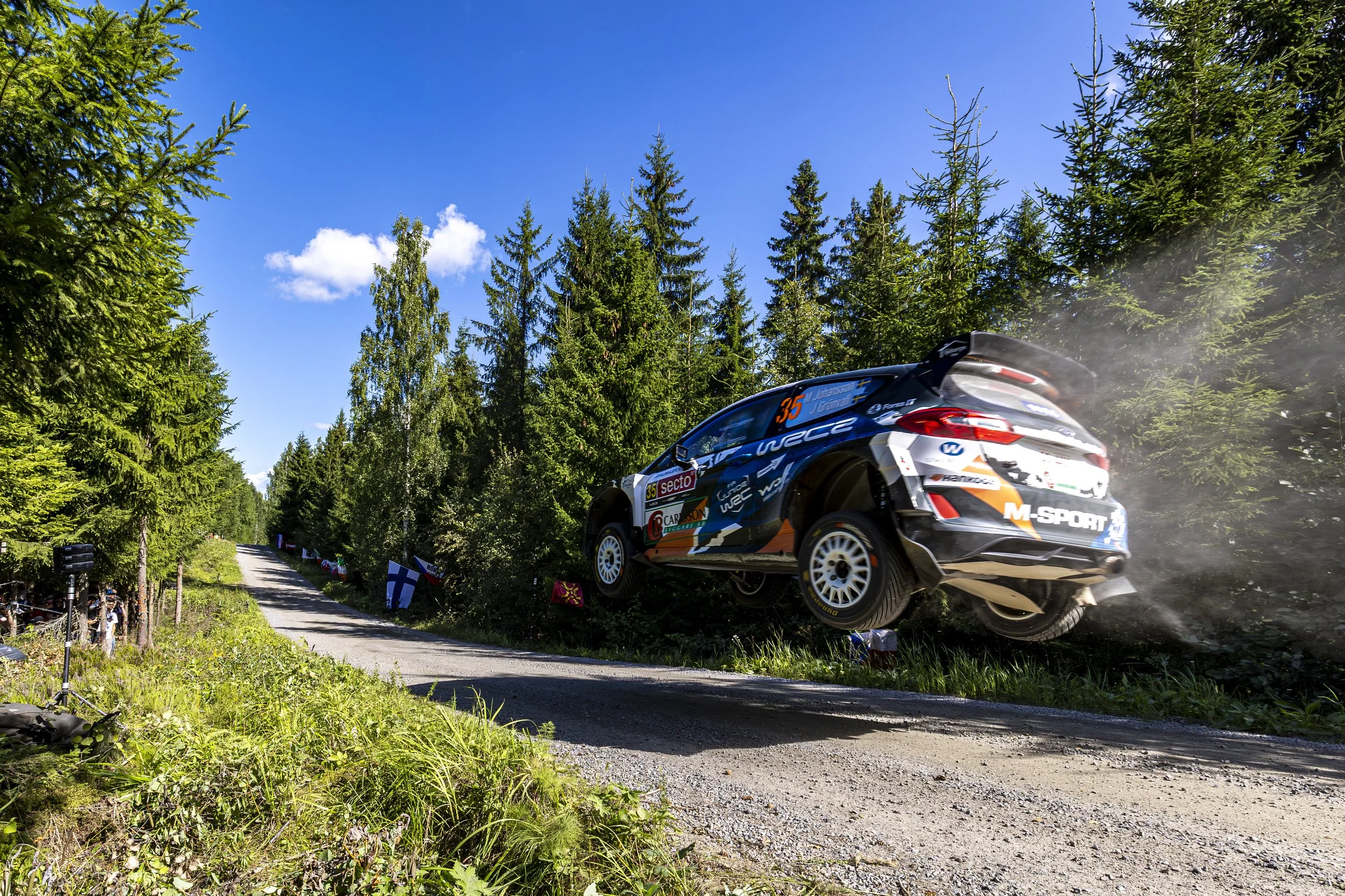 A rally car in mid-air jumping over a dirt road surrounded by tall evergreen trees under a blue sky.