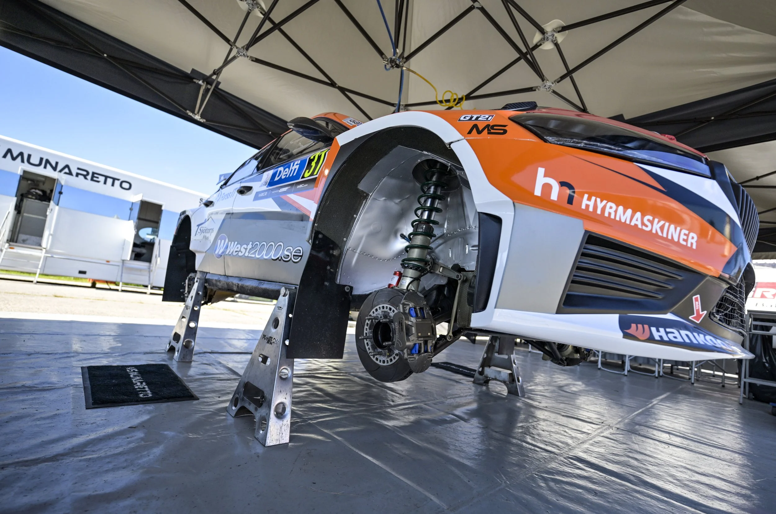 A racing car on a stand under a canopy at a race track, with sponsors and number 37 visible on the side.