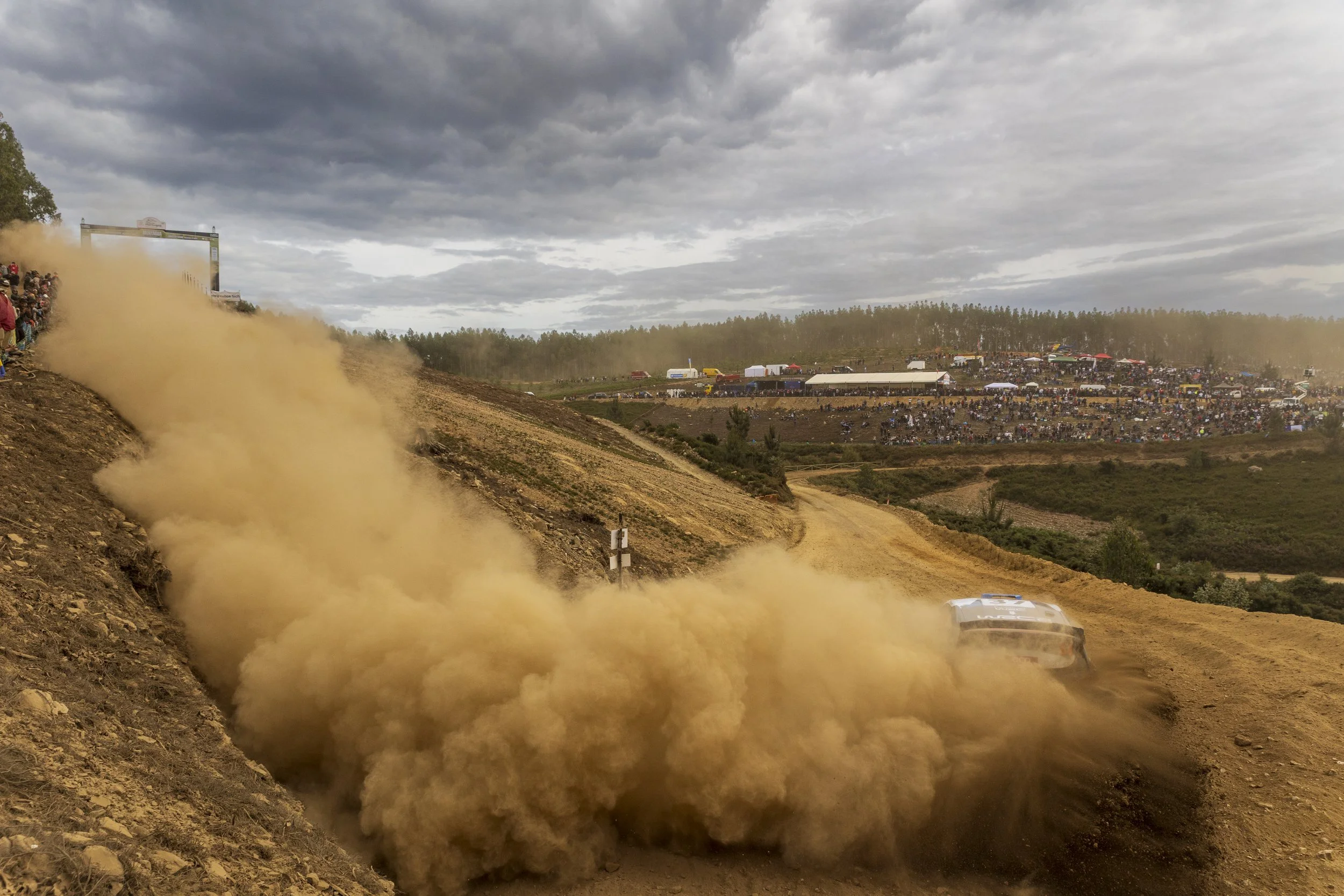 A rally car racing on a dirt track raising a large cloud of dust, with a crowded spectator stand in the background under a cloudy sky.