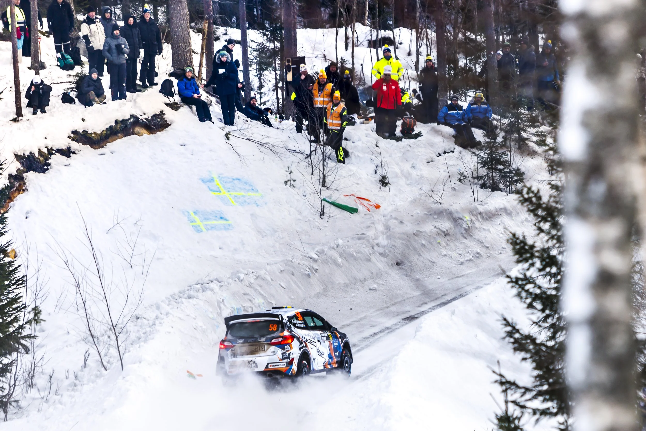 A rally car racing on a snow-covered mountain road during a winter event, with spectators and officials watching from the roadside.