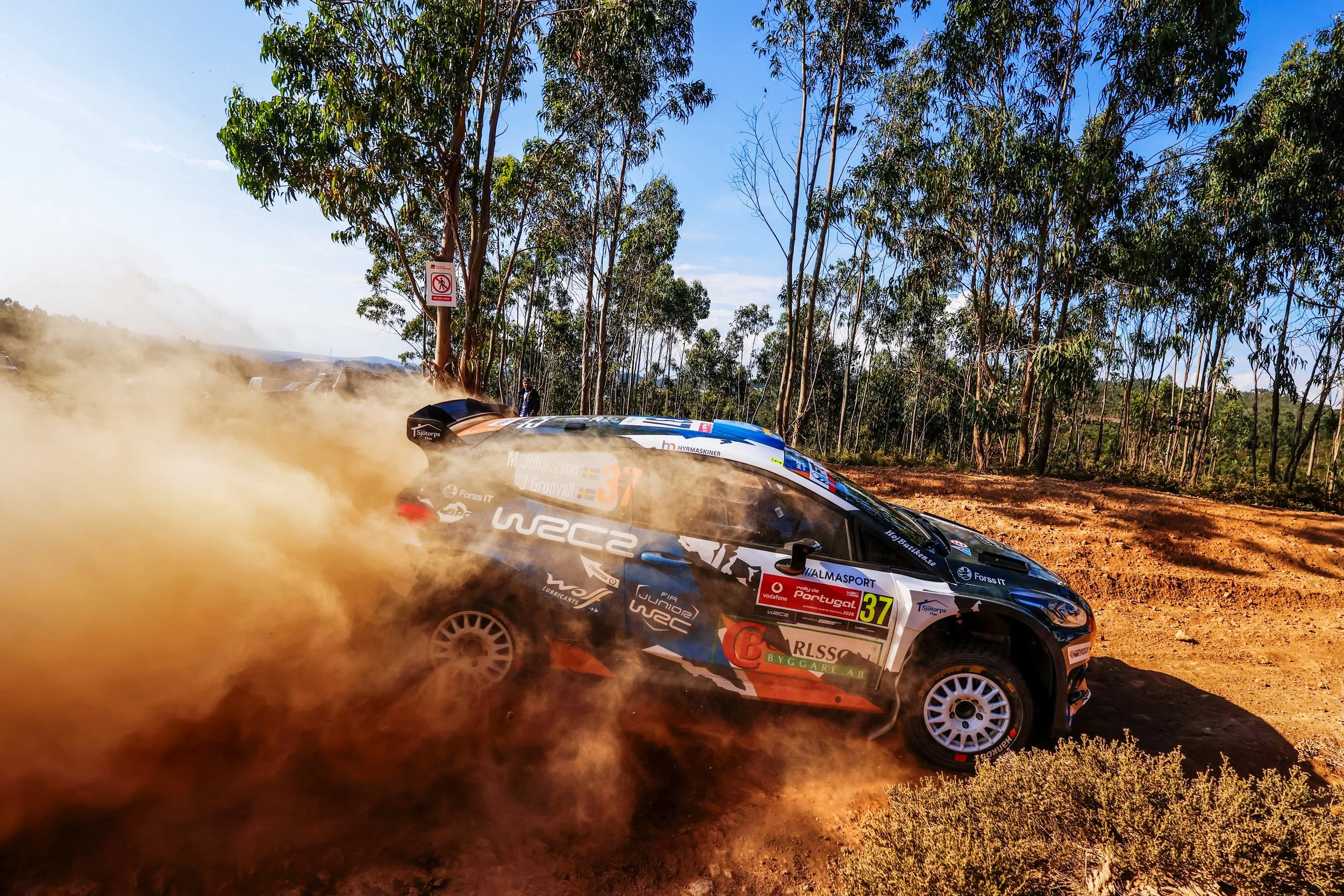 A rally car kicking up dust on a dirt road surrounded by trees under a blue sky.