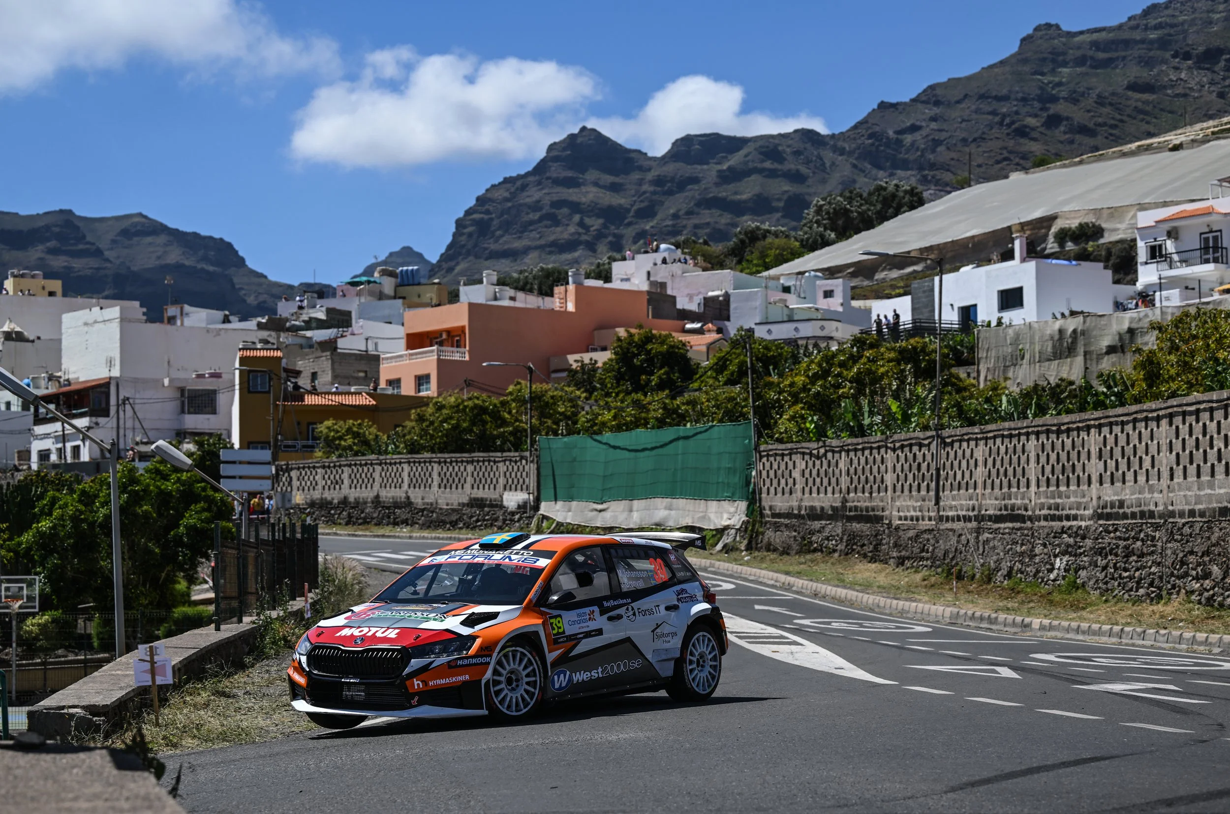 A rally car driving on a curving road in a mountainous area with houses on a hillside and a blue sky with clouds in the background.