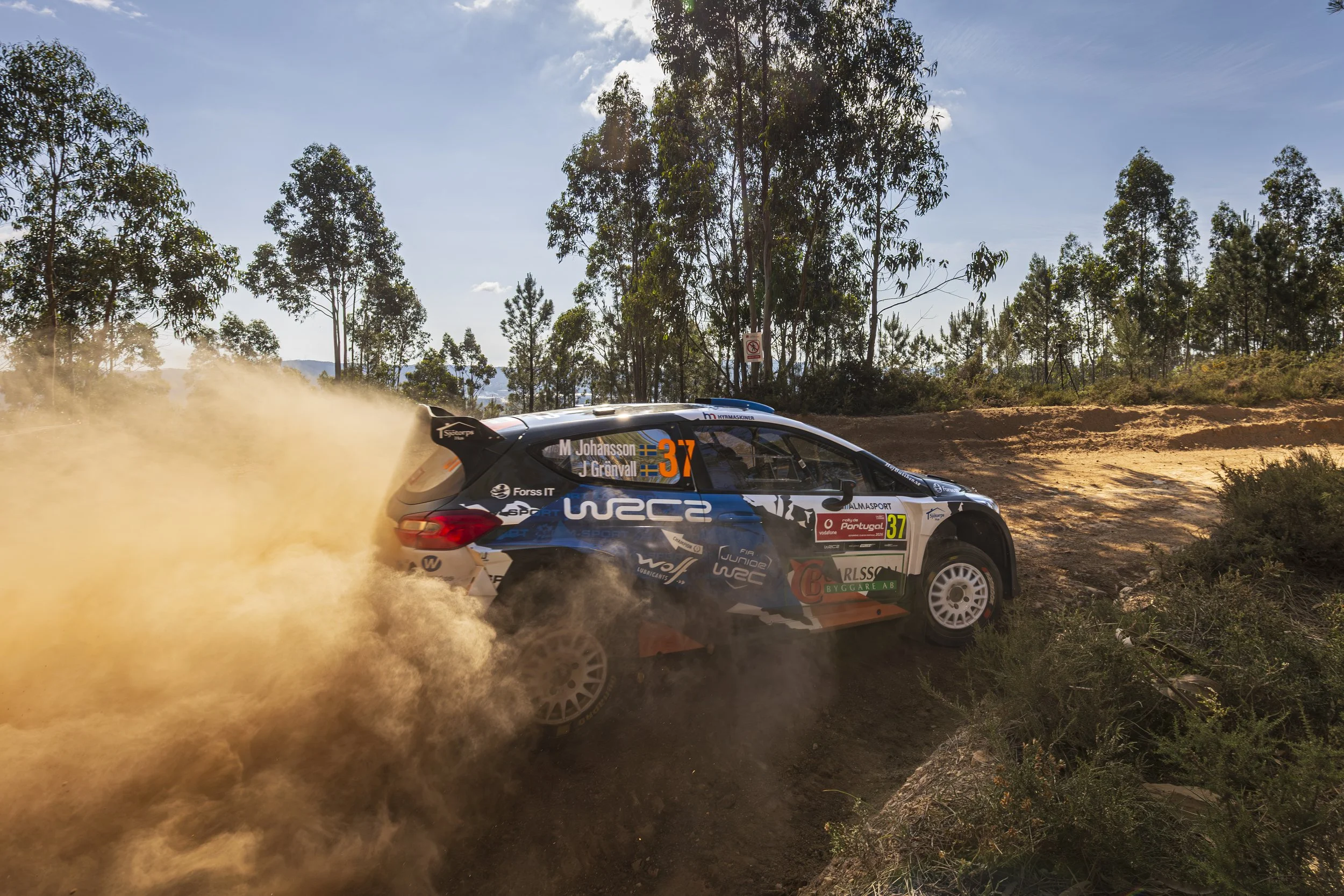 A rally car speeds through a dirt track, kicking up dust behind it as it navigates a turn surrounded by trees and foliage on a sunny day.