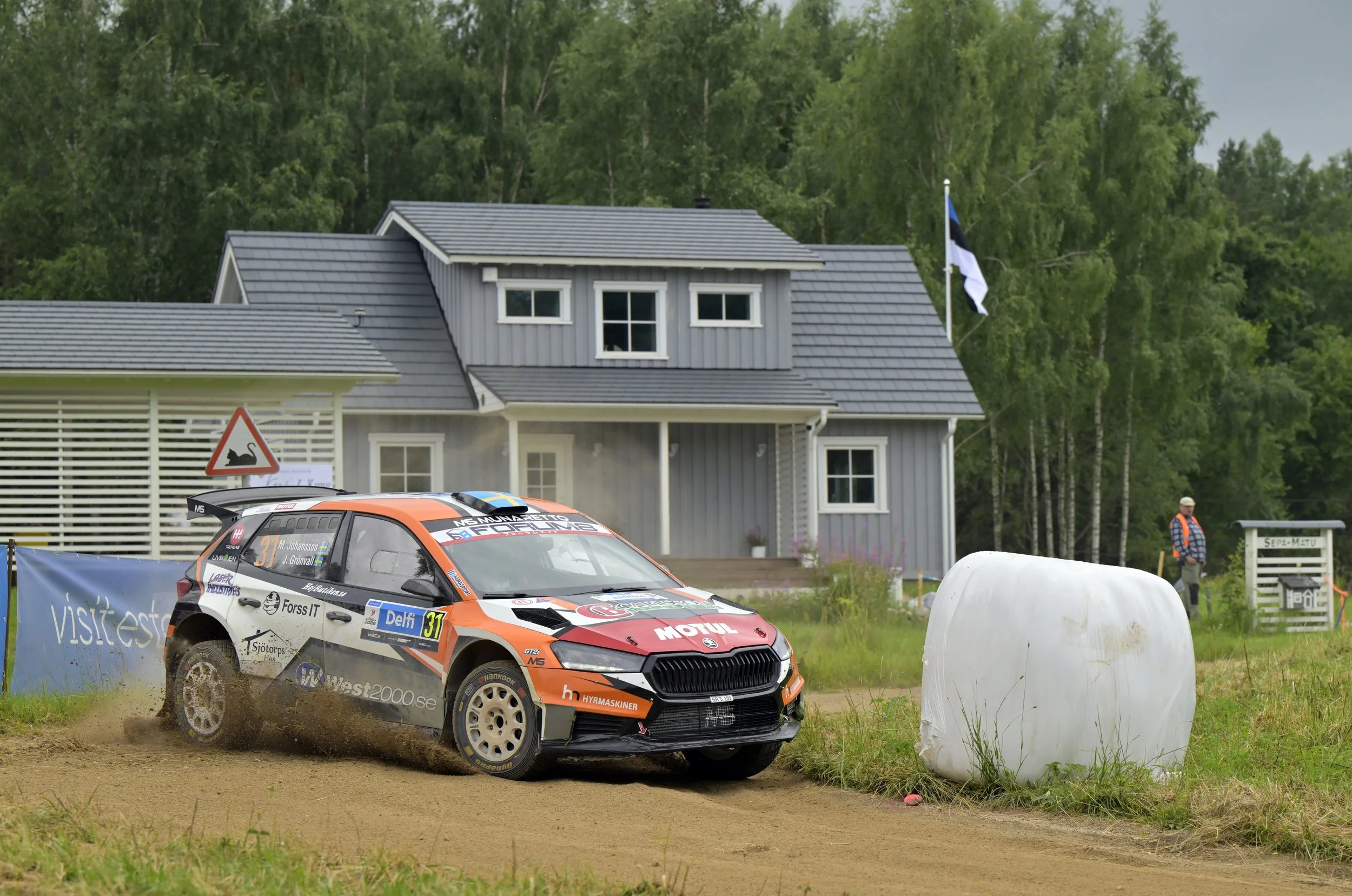 A rally car racing on a dirt track with dust flying behind it, next to a white hay bale and a man in a safety vest watching. In the background, there is a house with a gray roof and a flagpole with a blue, black, and white flag, green trees, and a bl