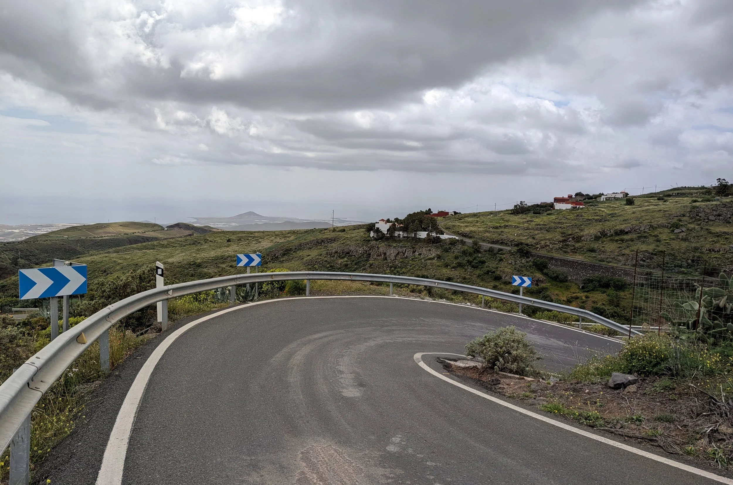 A winding mountain road with guardrails and road signs, overlooking a hilly landscape with scattered houses and cloudy sky.