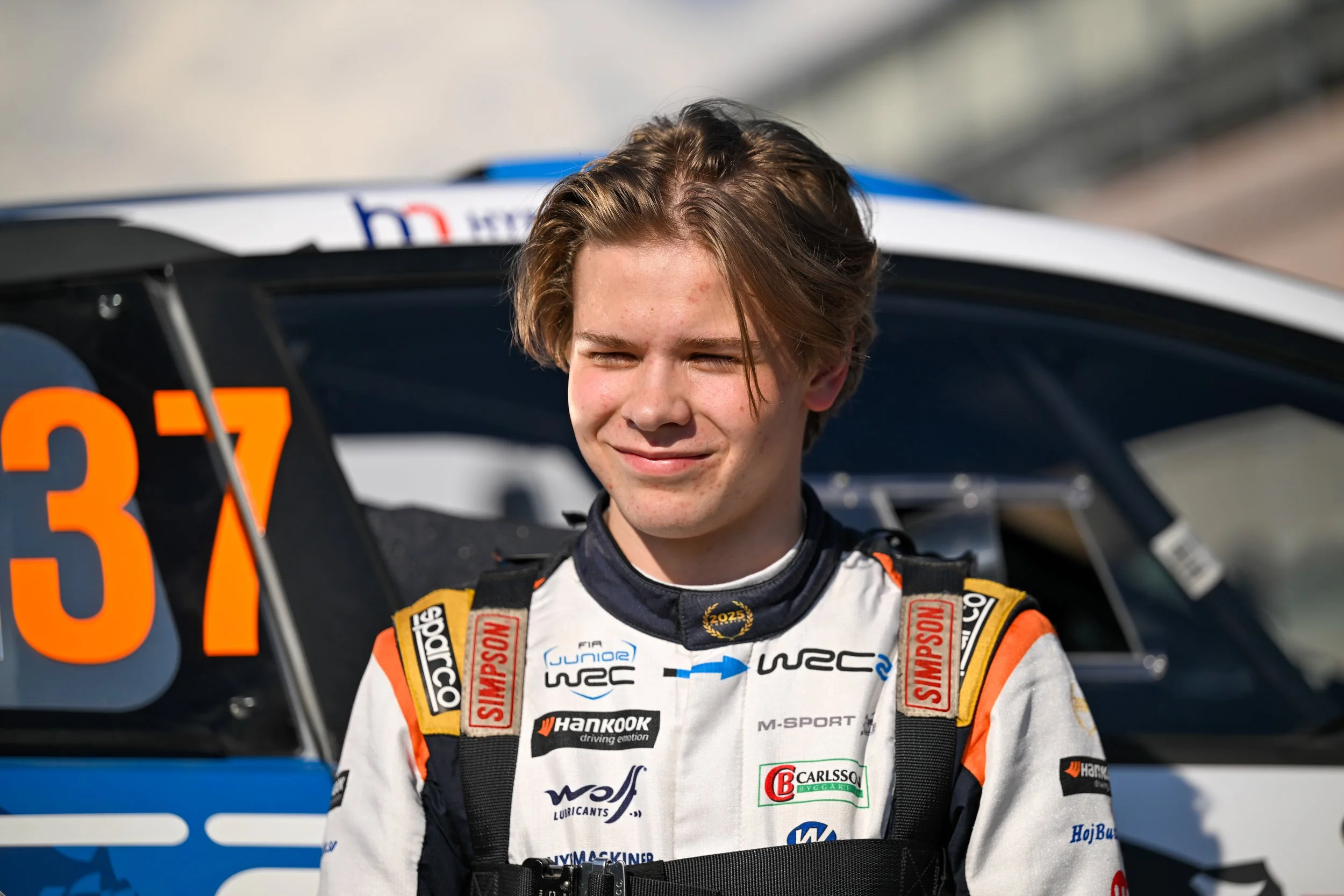 A young male race car driver in a racing suit with sponsor logos, standing in front of a race car. He is smiling with eyes closed, and his hair is tousled.