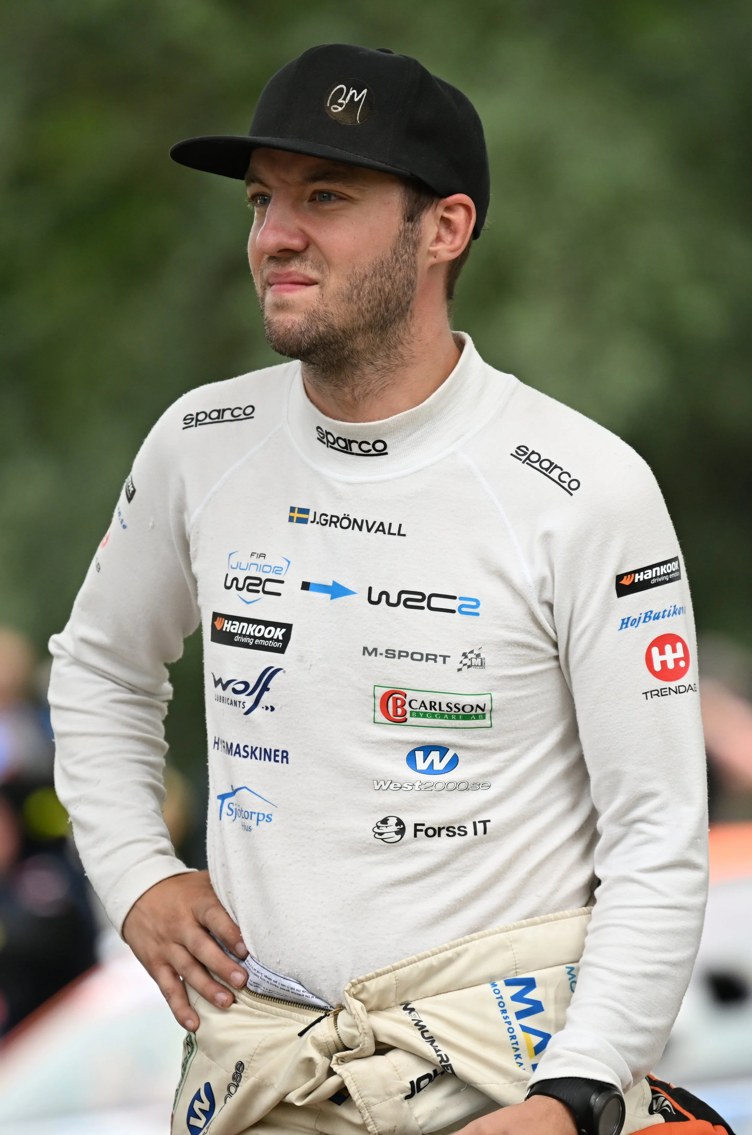 A male racing driver standing outdoors, wearing a white racing suit with sponsorship decals, a black cap with a logo, and beige racing pants with more sponsor logos. He has light skin, a beard, and short hair, with one hand on his hip and looking to 