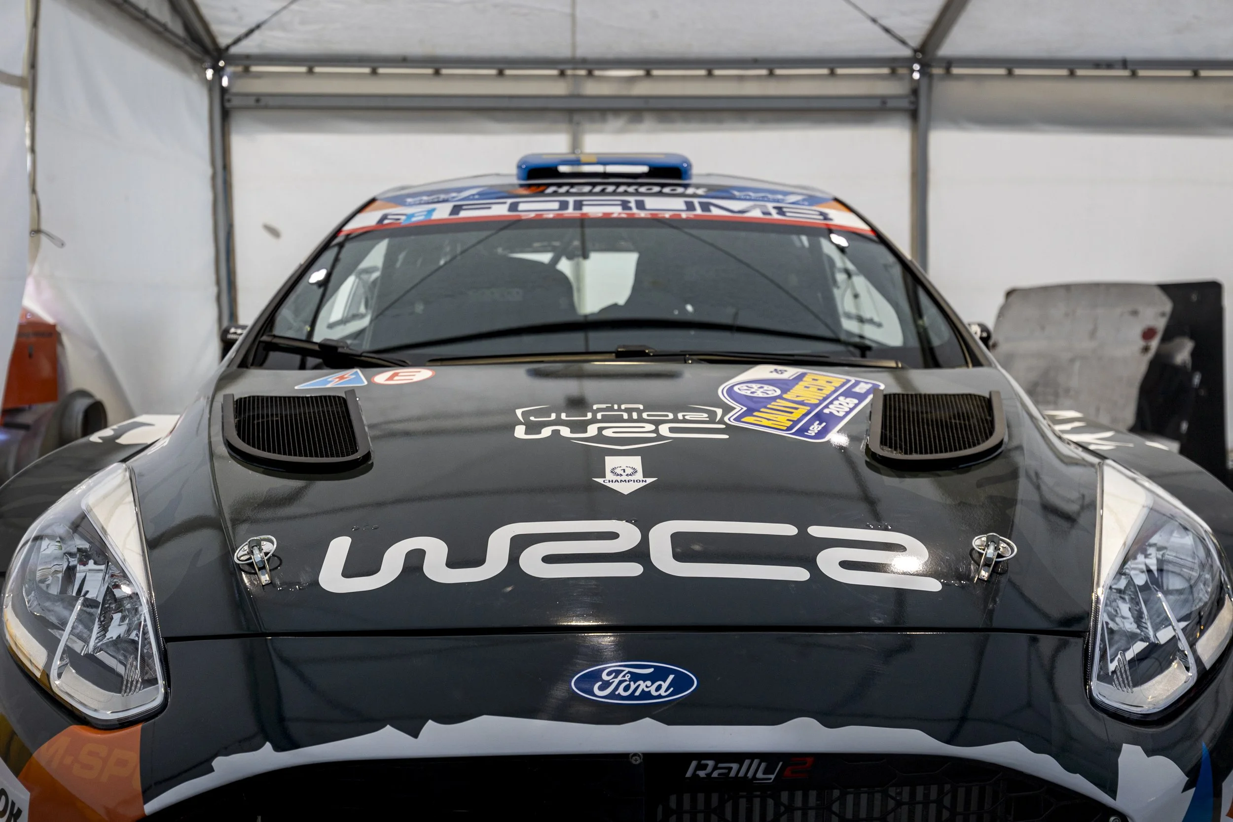 A black rally car with Ford logo on the front, featuring racing stickers and decals, parked inside a garage or tent.