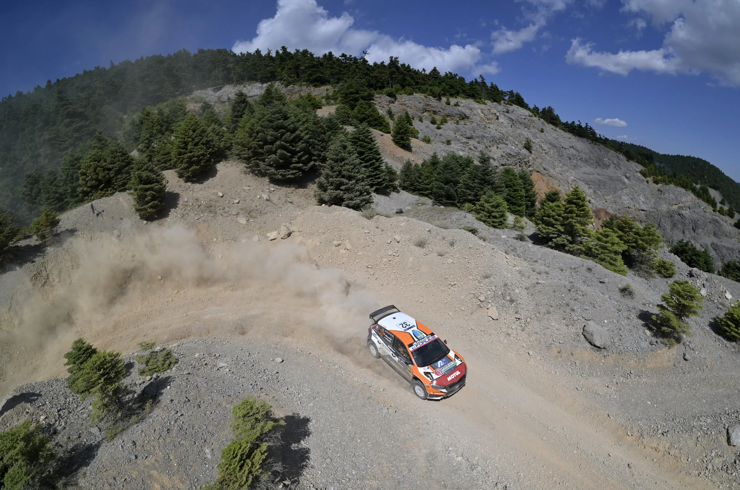 A rally car racing on a dirt mountain road, kicking up dust, surrounded by pine trees and rocky terrain under a partly cloudy sky.