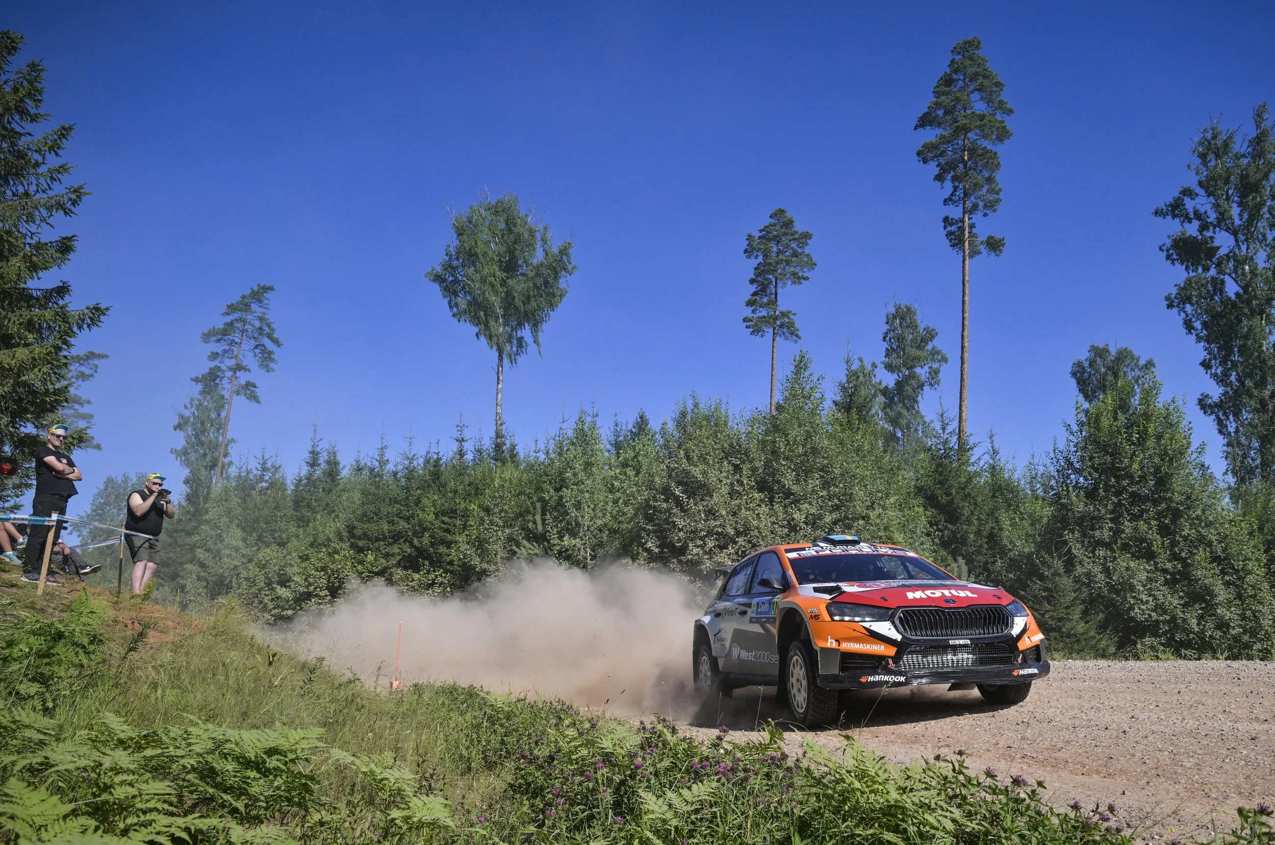 A rally car racing on a dirt road in a forested area, kicking up dust, with spectators watching from the side of the track during a sunny day.