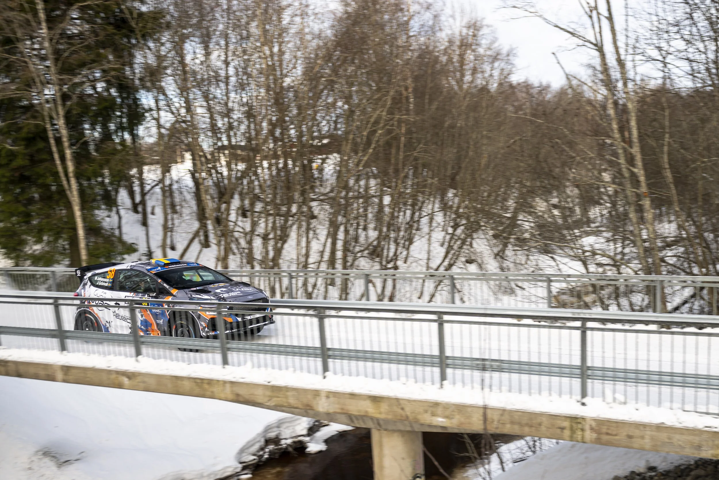 A rally car racing on a snowy road, with leafless trees in the background.