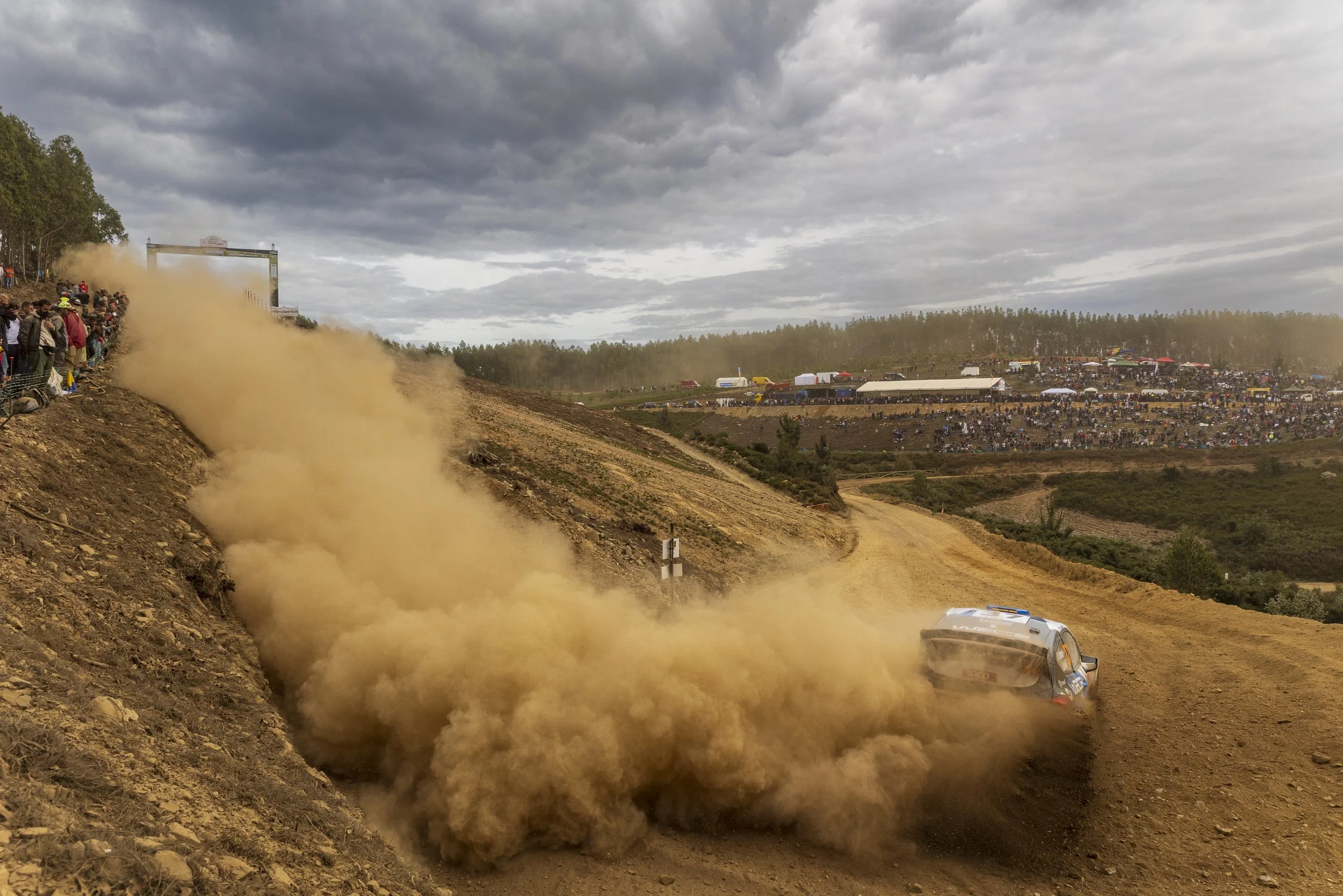 A dirt racetrack with a crowd of spectators on the left and in the background, motorhomes, trailers, and tents. A race car is sliding off the track, kicking up a large cloud of dust under a cloudy sky.