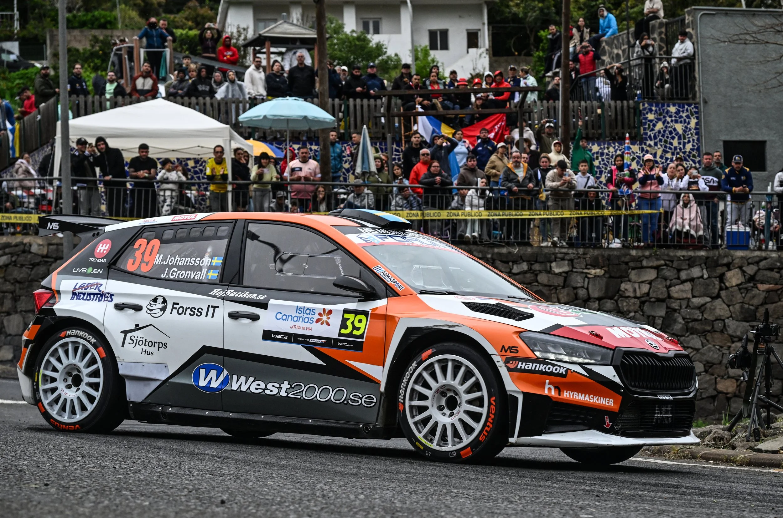 A rally car with sponsors and racing numbers parked on a wet road, with a crowd of spectators watching behind a fence in the background.