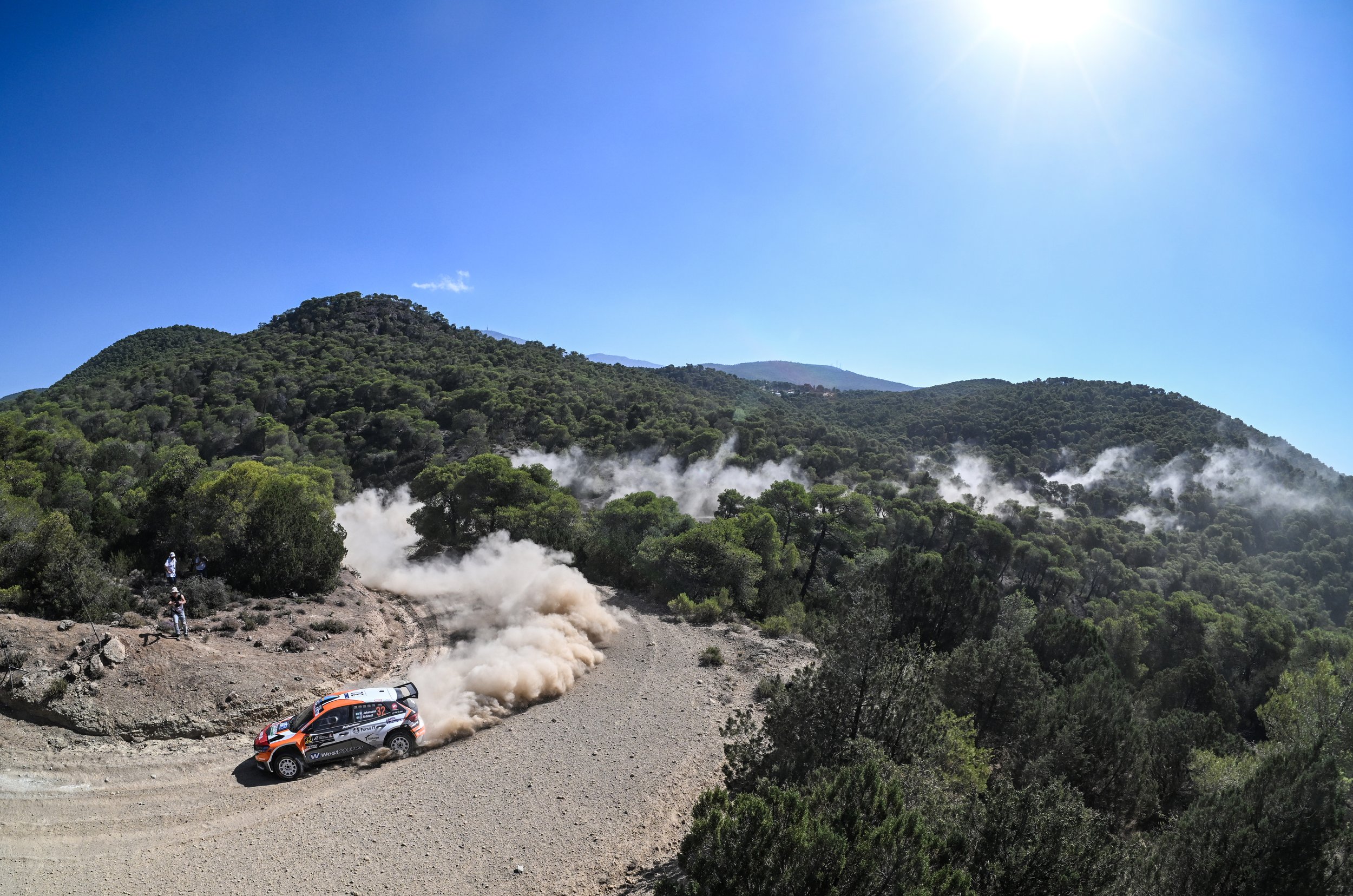 A rally car racing on a dirt mountain road, kicking up dust as it speeds by in a forested area with hills and mountains in the background under a clear blue sky.
