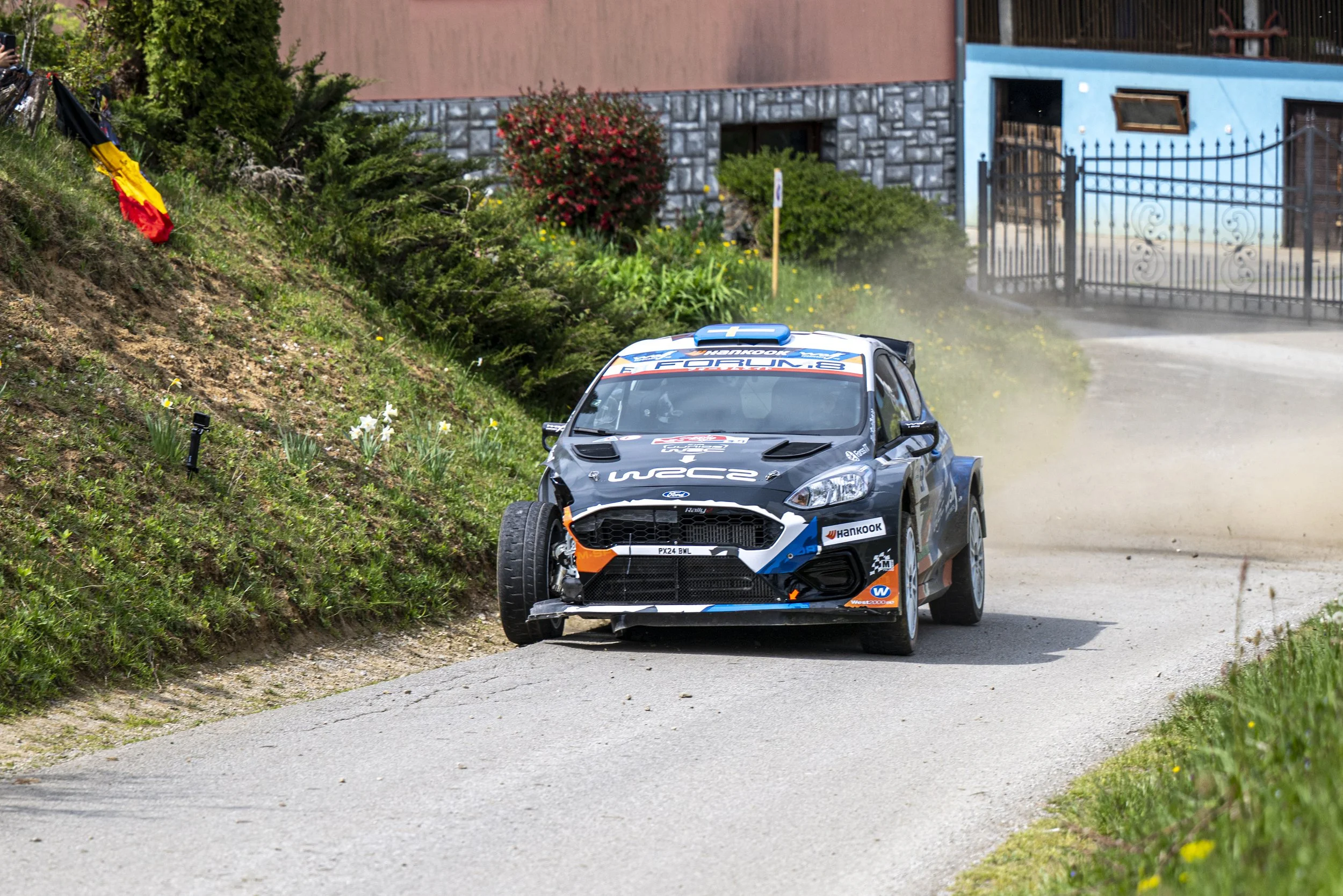 A rally car driving on a dirt road next to a grassy hillside with flowers, with a house and gate in the background.