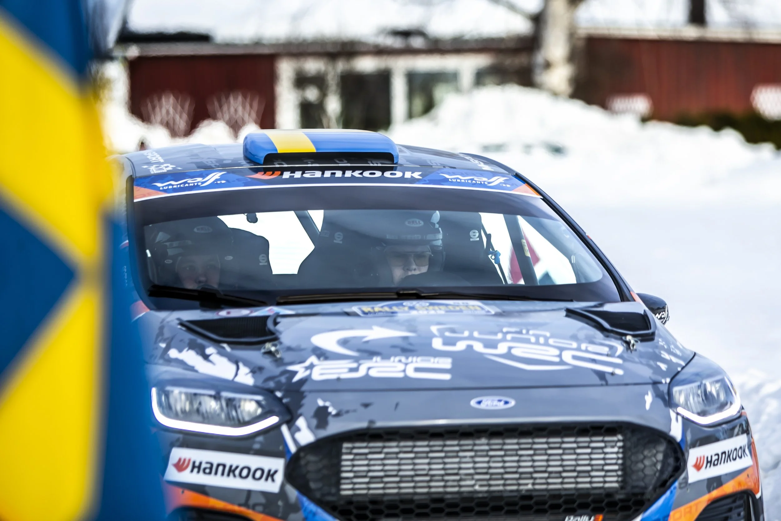 Close-up of a black rally car with Hankook sponsorship, driven by racers wearing helmets, on snow-covered terrain during a race, with a red building and trees in the background.