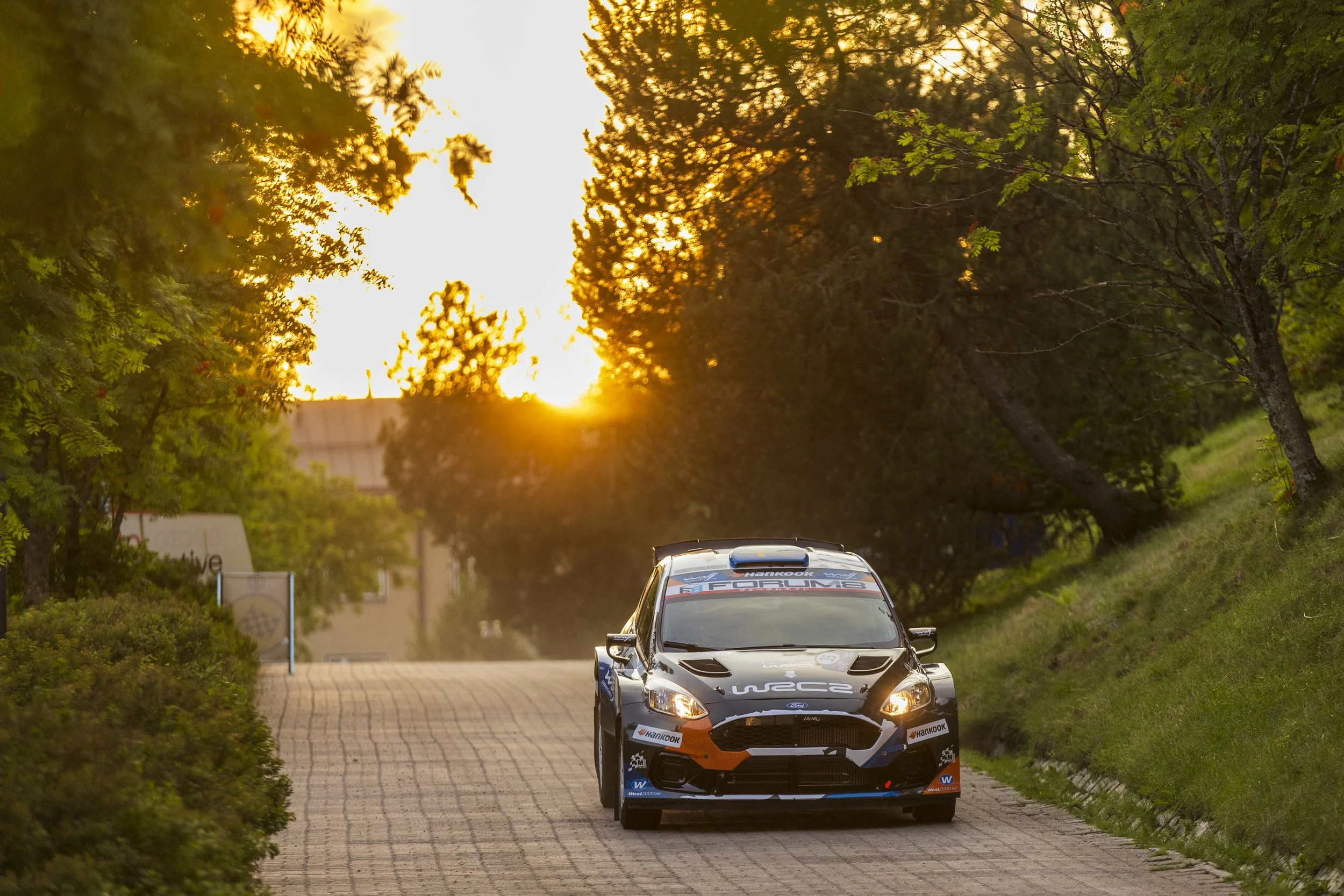 A rally car driving on a narrow paved road surrounded by trees during sunset.