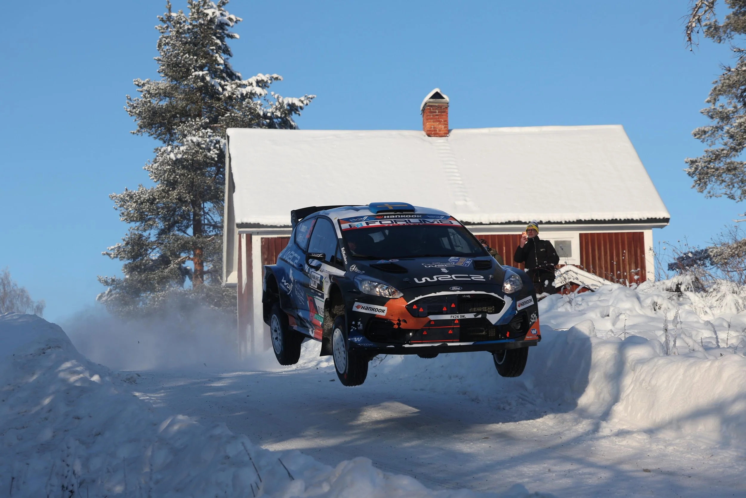 A rally car is airborne above snowy ground during a race, with a person standing next to a wooden house and snow-covered trees in the background on a clear winter day.