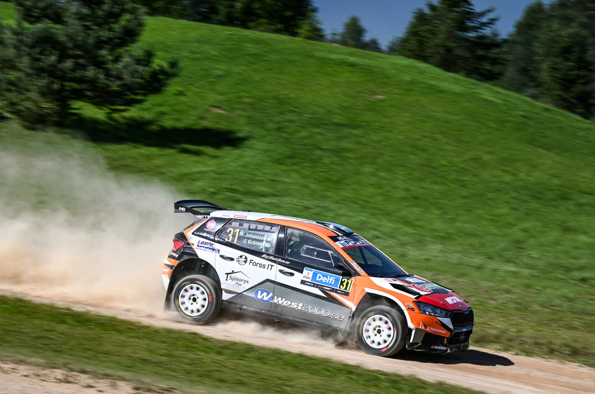 A rally car racing on a dirt trail with a grassy hill and trees in the background during the daytime.