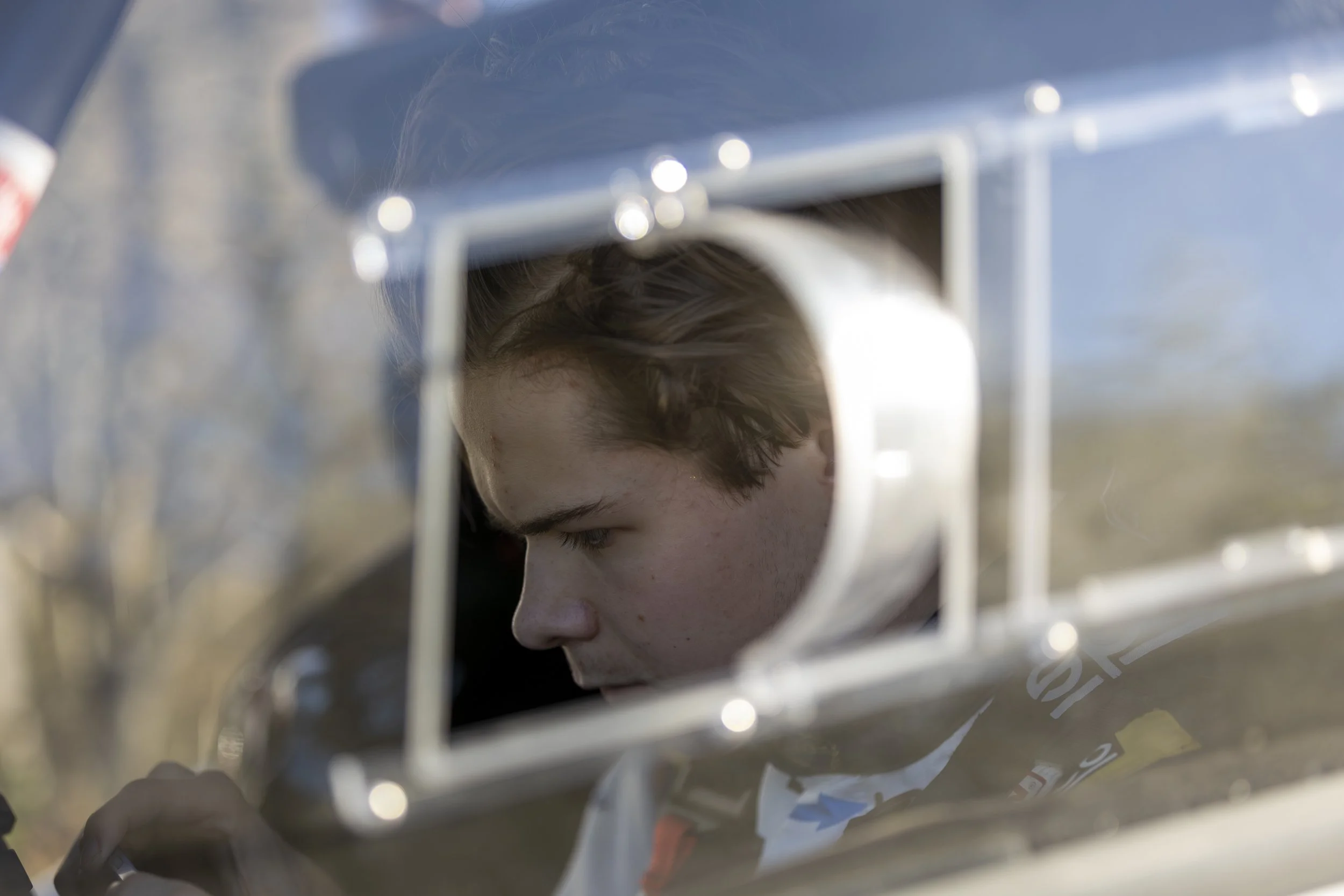 A person's face with short, dark hair, viewed through the bars of a metal cage or enclosure, outdoors with a clear sky in the background.
