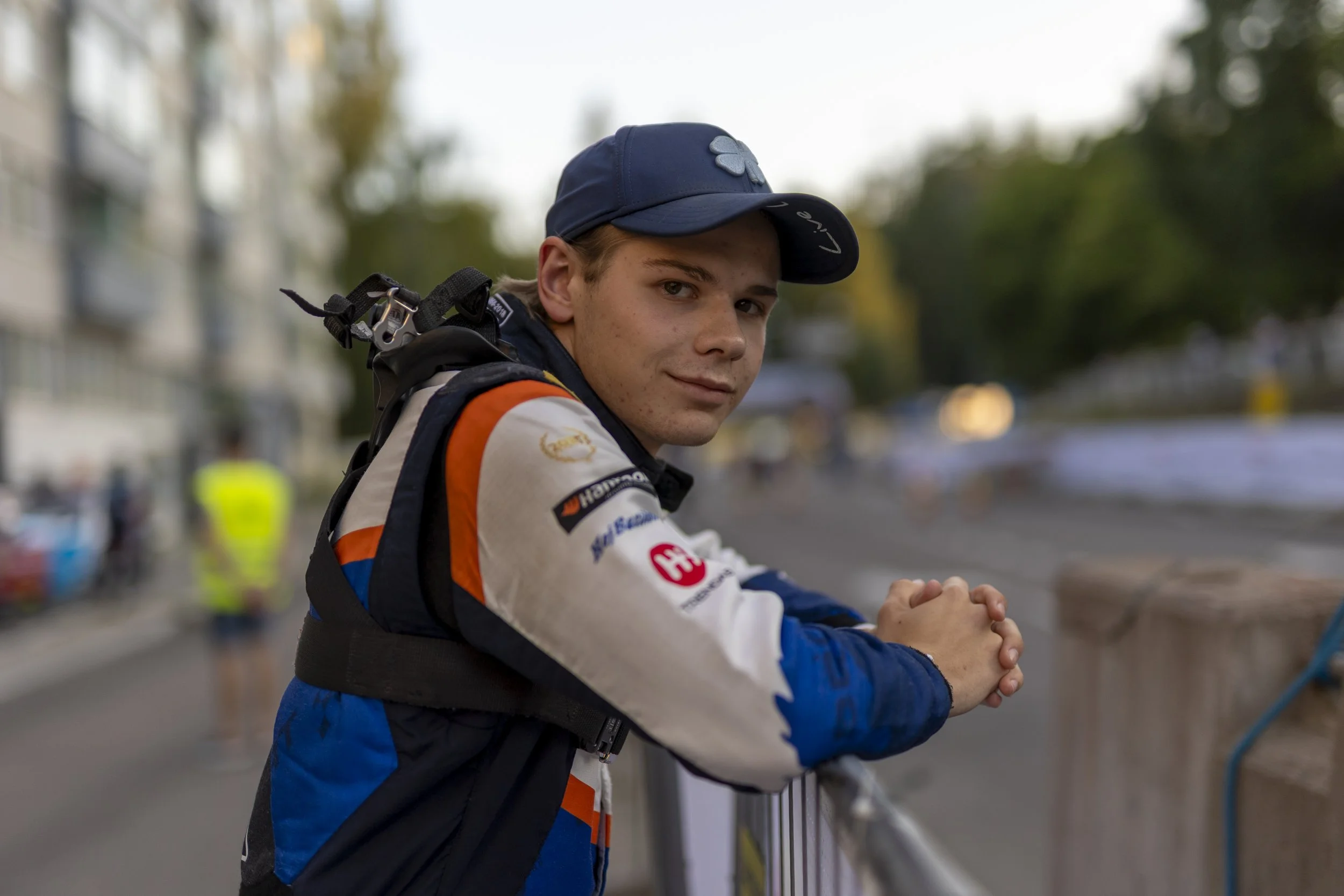 A young male race car driver wearing a racing suit and cap, leaning on a fence with a smile, outdoors at a racing event.
