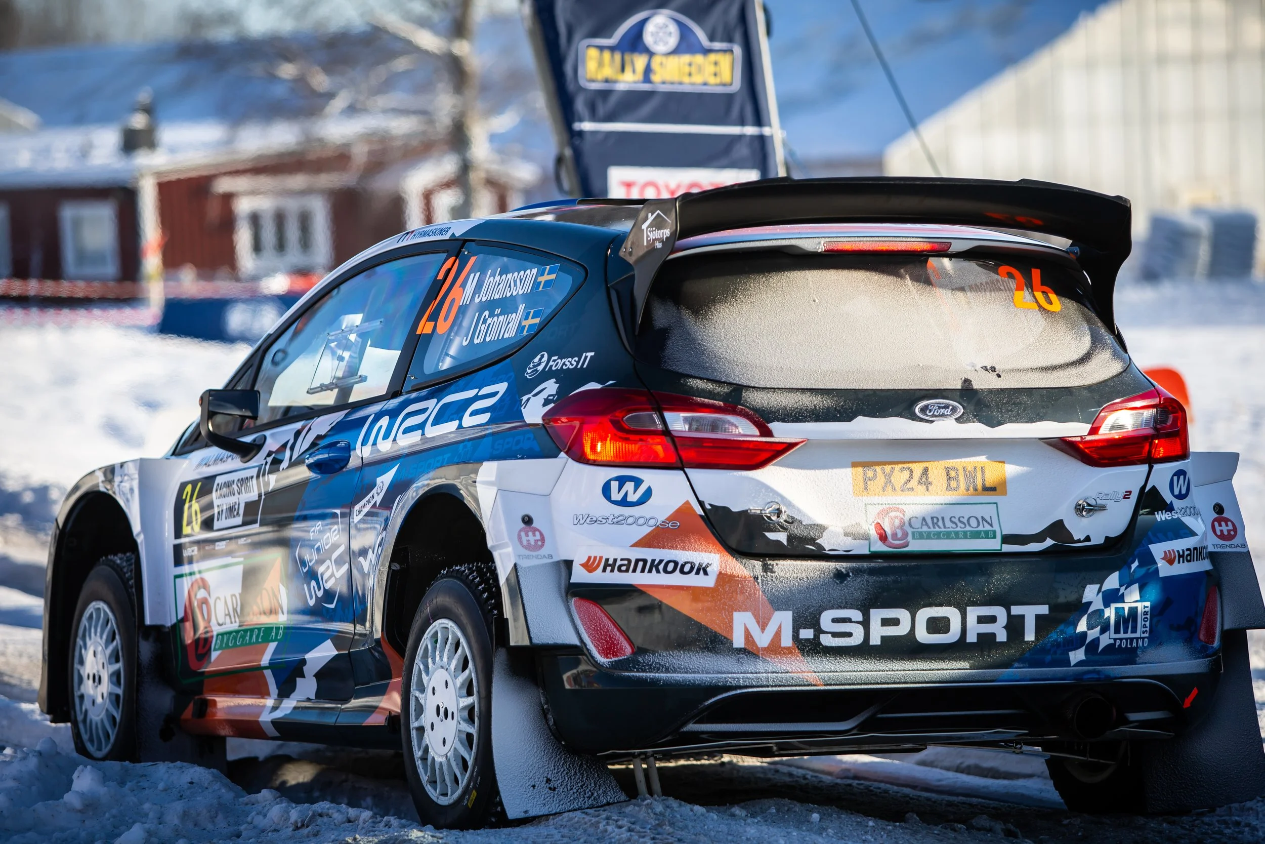 A rally car on snow-covered ground with sponsor logos and the number 26, driven by M. Johansson and J. Grönvall, in a winter setting with a house and trees in the background.