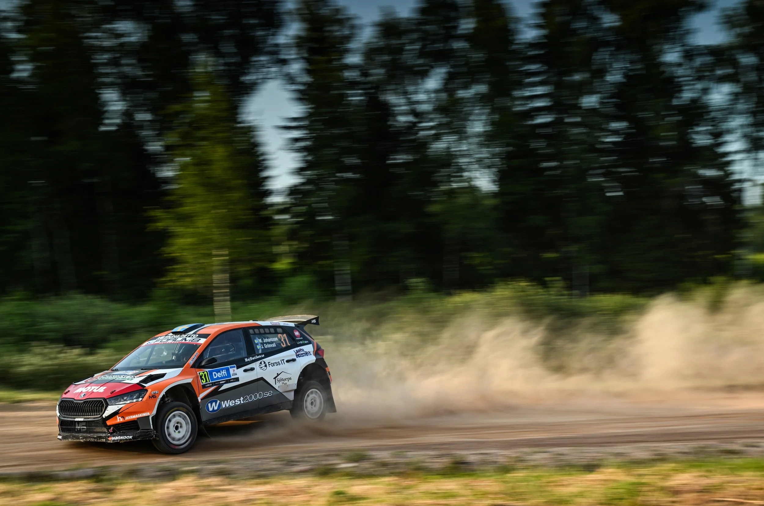 A rally car racing along a dirt road, kicking up dust with a blurred forest background.