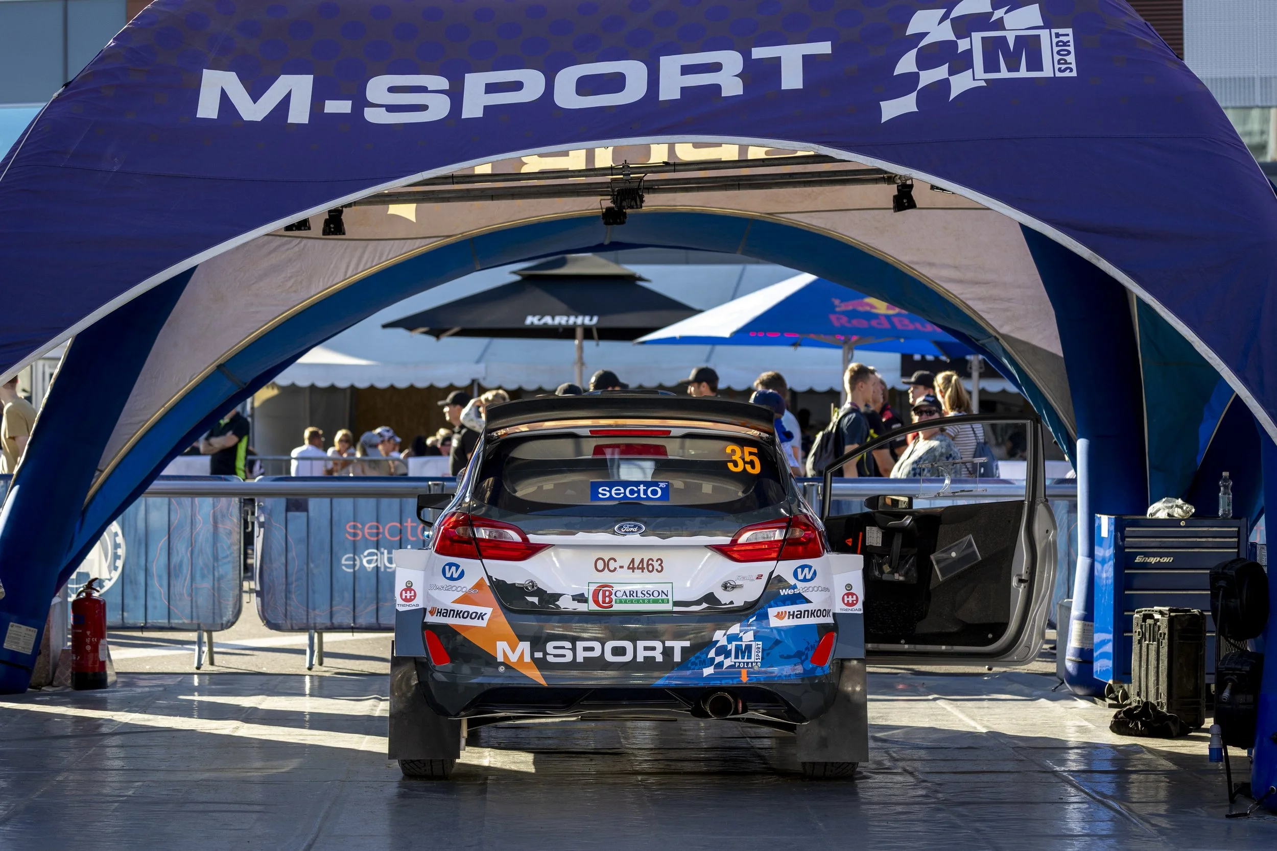 Race car parked in the pit lane under a blue tent with M-Sport branding, spectators and team members in the background.