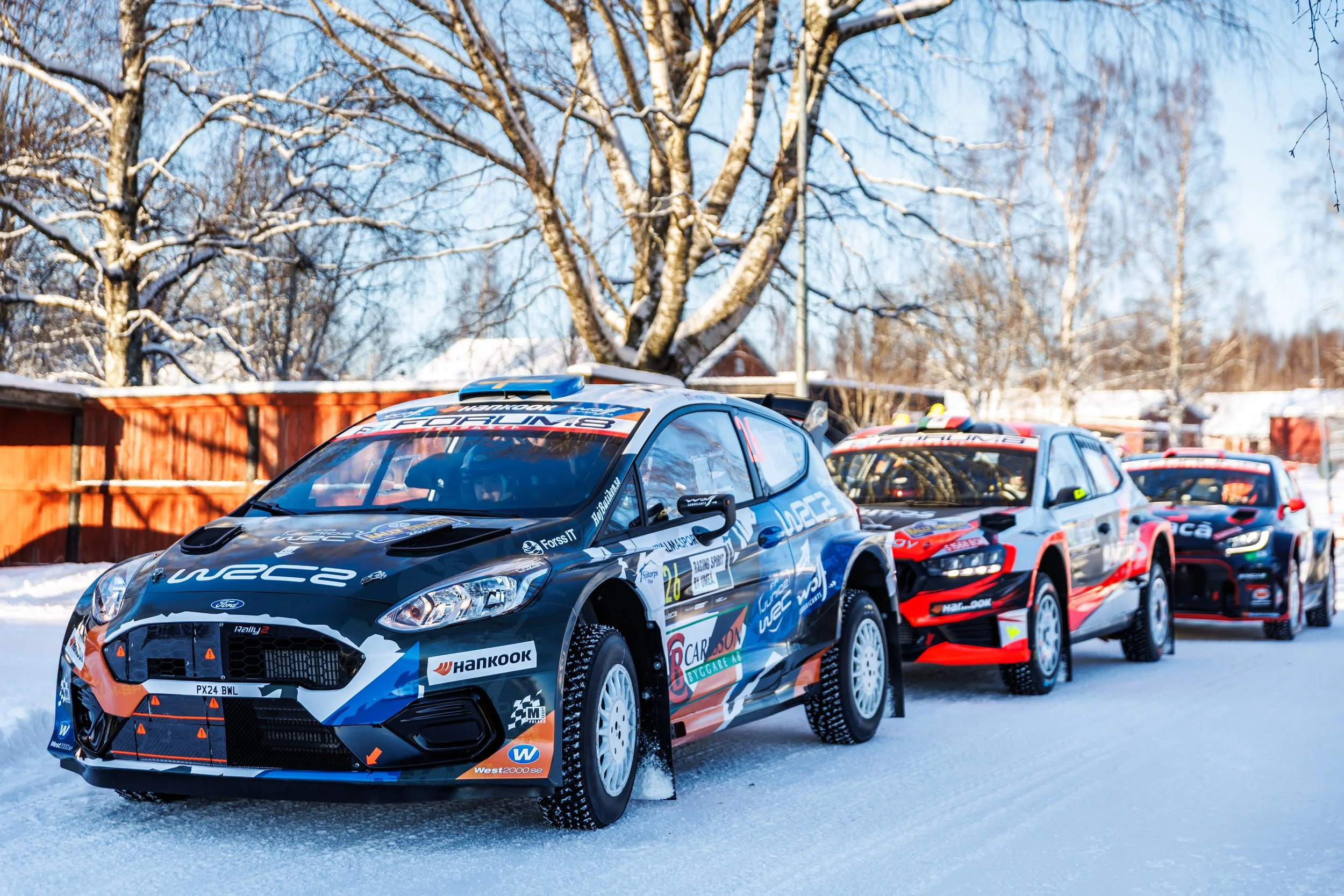 Rally cars racing on a snowy track with snow-covered trees and a wooden fence in the background.