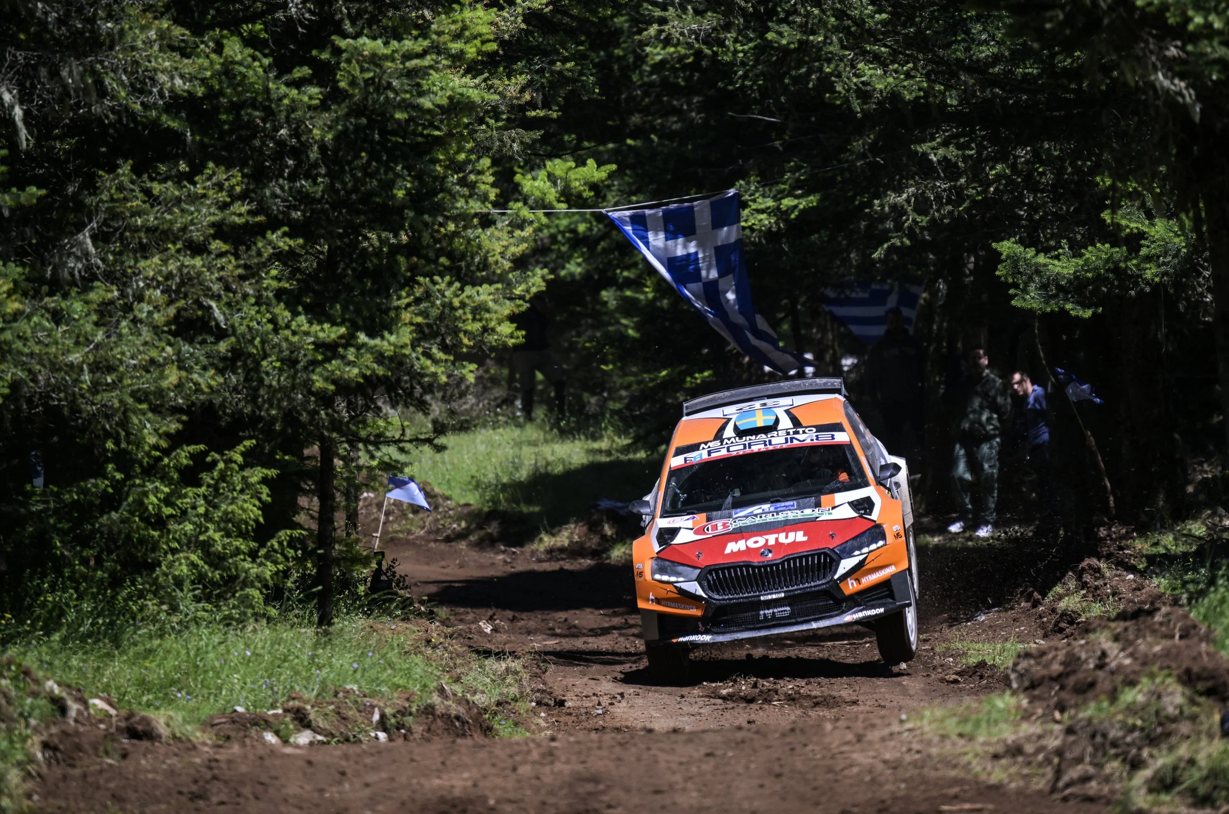 An orange rally car racing on a dirt trail through a forest with trees and greenery, with flags hanging across the track.