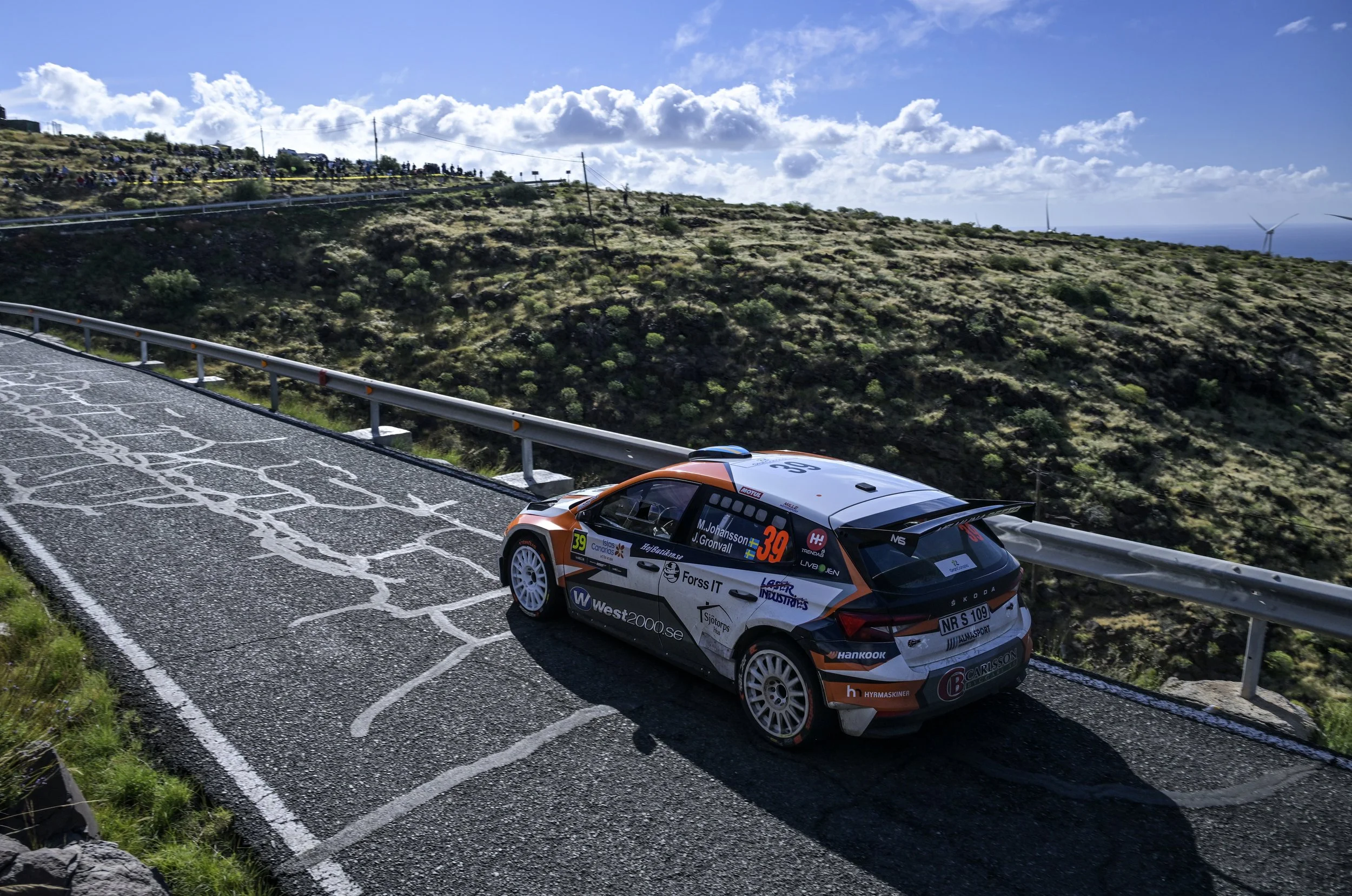 A rally car speeding on a mountain road with spectators watching from the hillside.