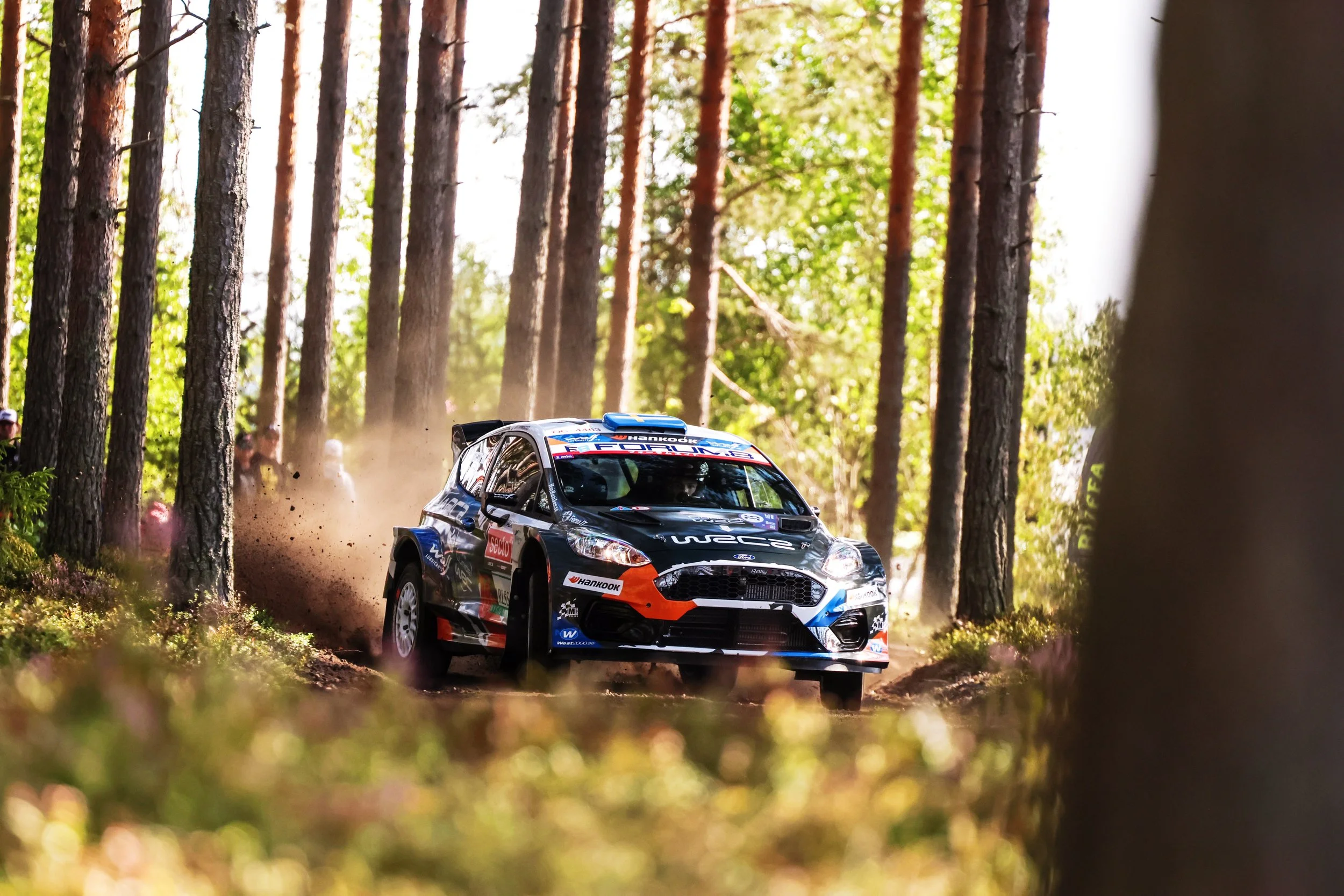A rally car driving through a forest trail surrounded by tall trees.