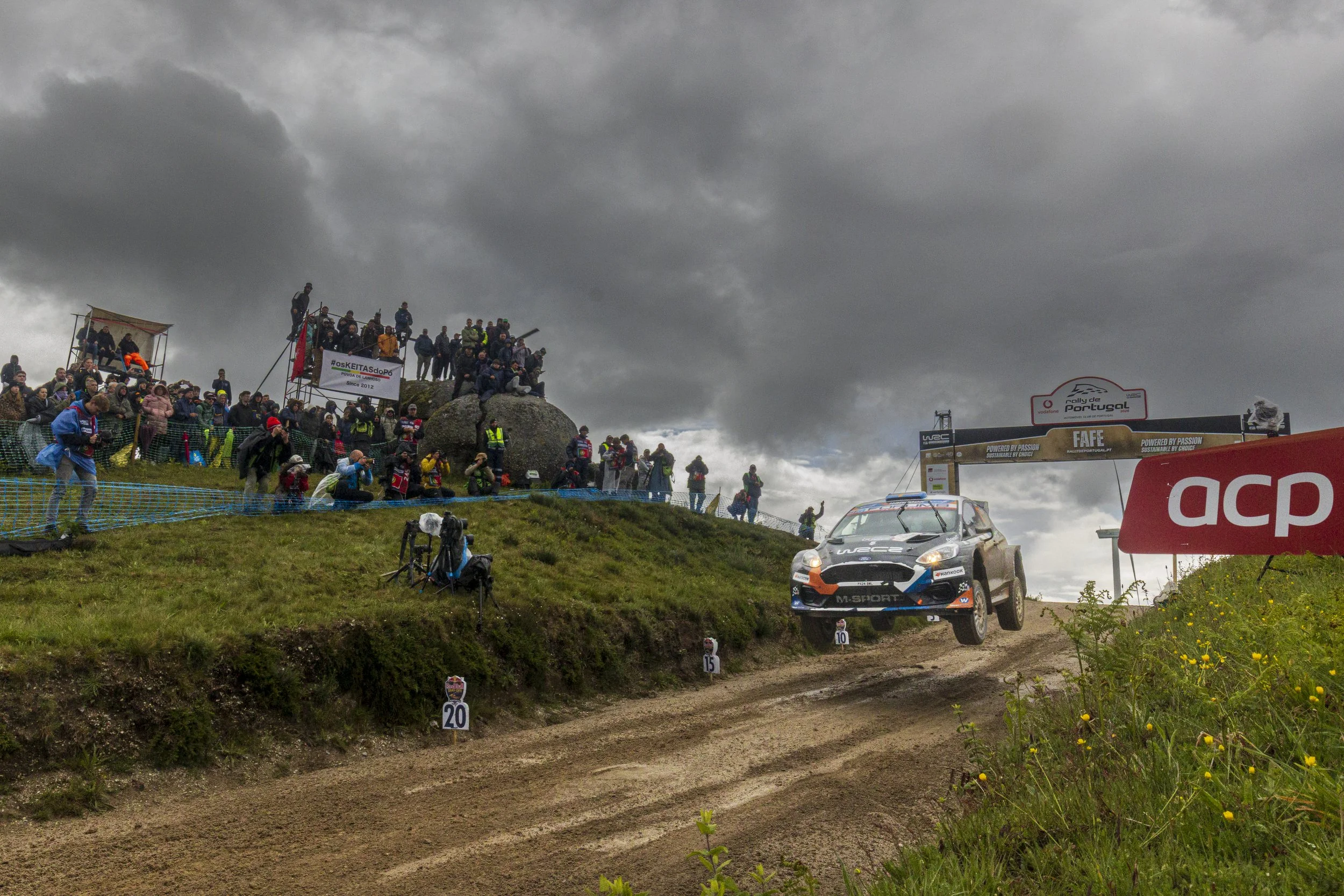 A rally car racing on a dirt track with a crowd of spectators watching from a hilltop, dark cloudy sky overhead, and rally signage visible.
