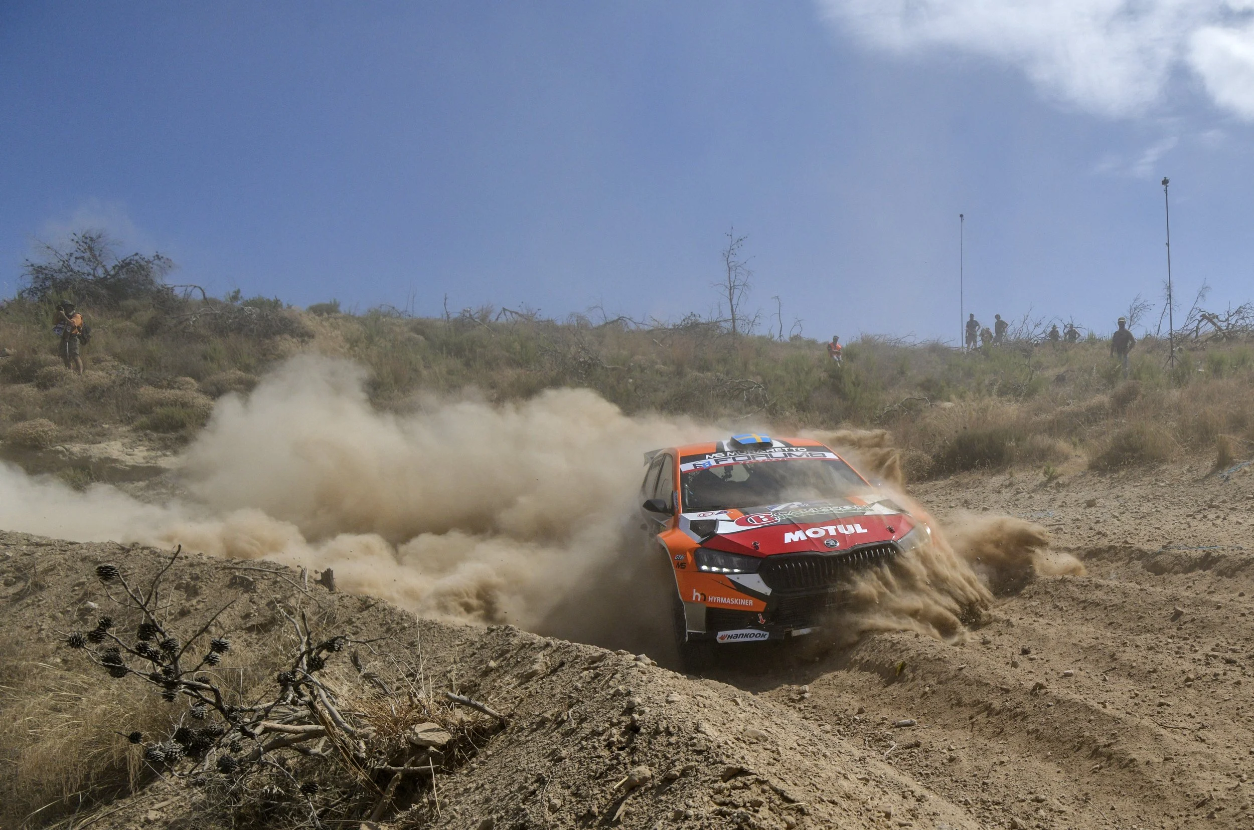 A rally car speeding on a dirt track, kicking up dust with spectators standing on a hill in the background under a blue sky.