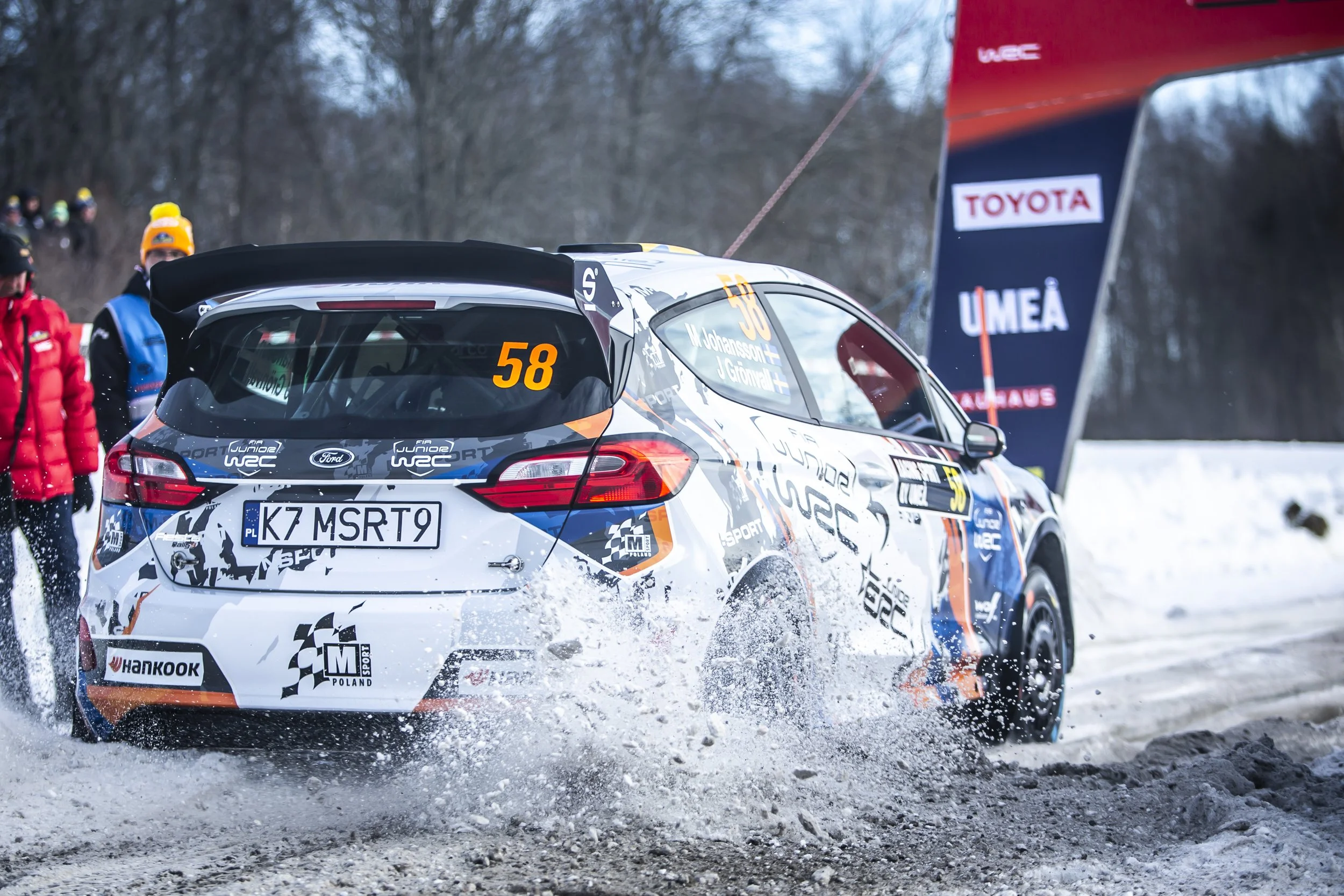 Rally car racing on snowy terrain, with a white car marked with the number 58 and sponsors, surrounded by snow spray and spectators nearby.