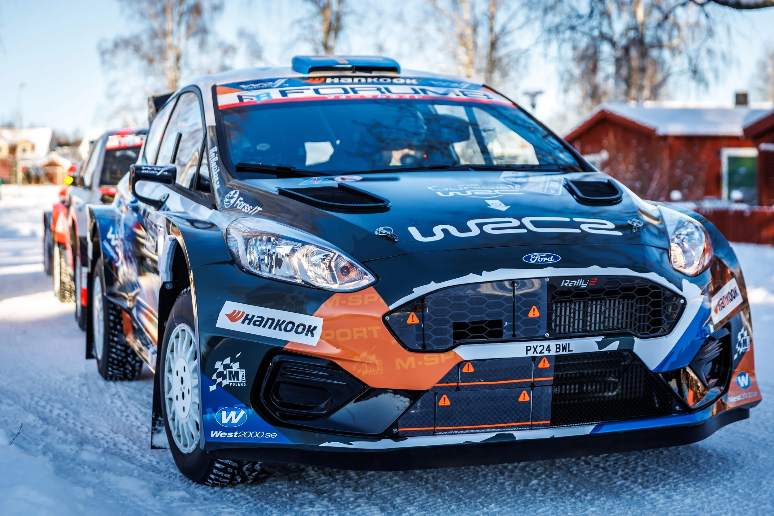 A black Ford rally car with sponsor logos, parked on snow, with trees and houses in the background.