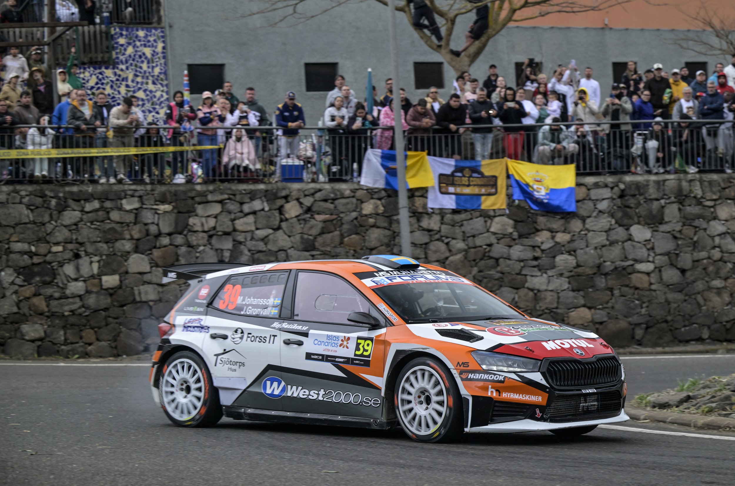A rally car racing on a city street with spectators watching behind a fence, banners, and a stone wall.