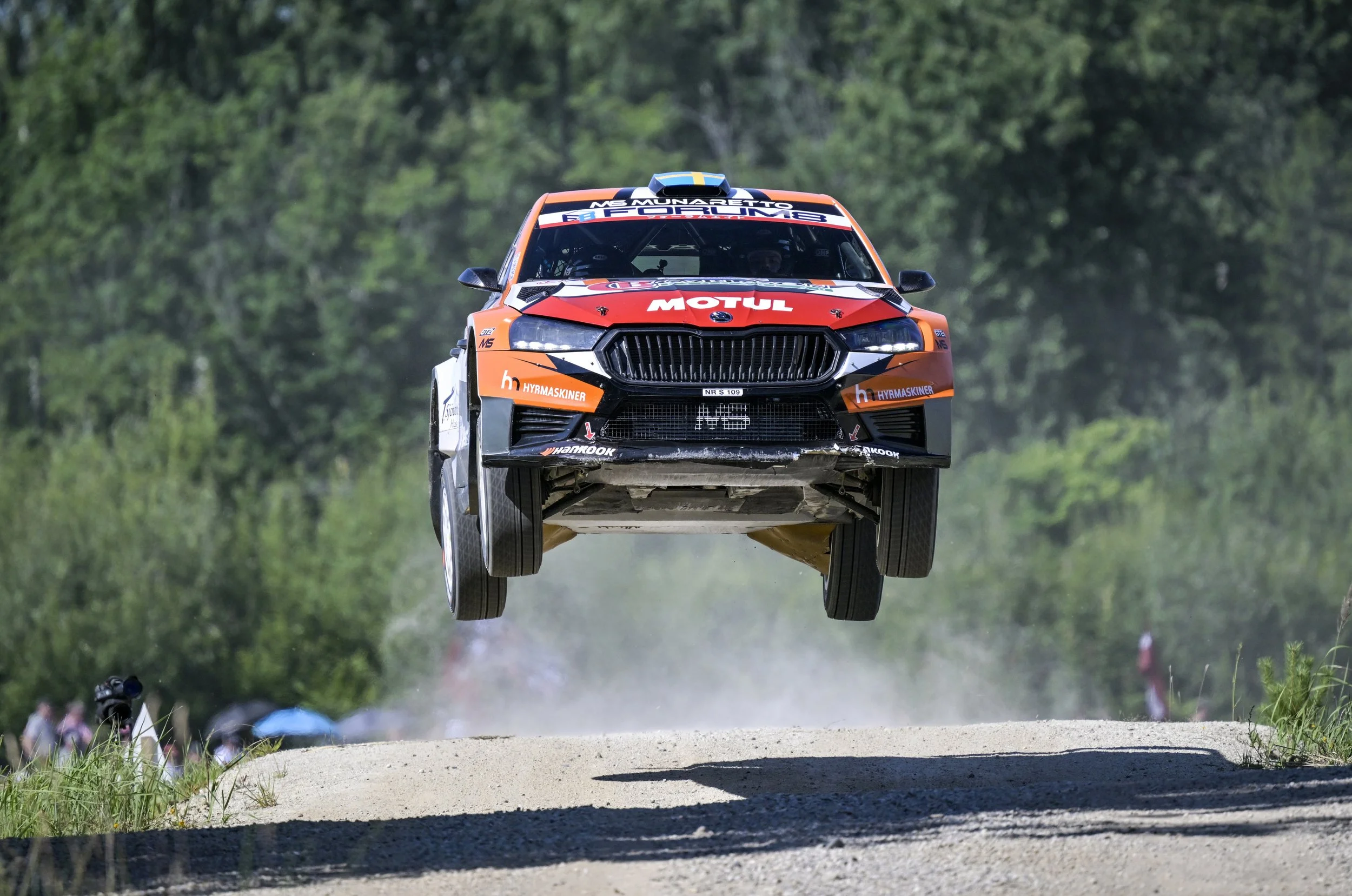 A rally car flying through the air over a dirt road in a forested area during a race.