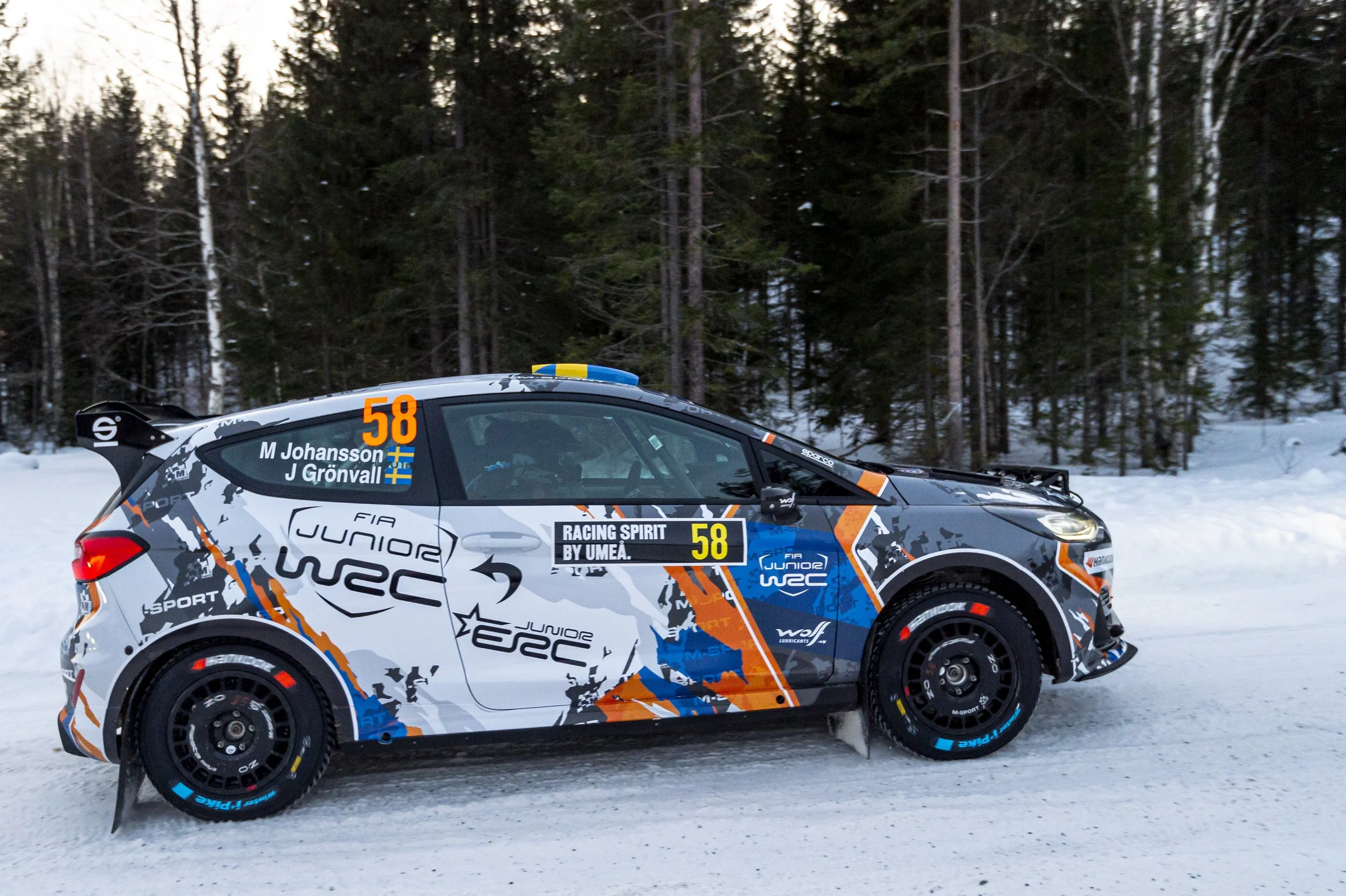 A rally car on a snowy forest road with sponsorship decals and the number 58, driven by M. Johansson and J. Grönvall, during a winter rally event.