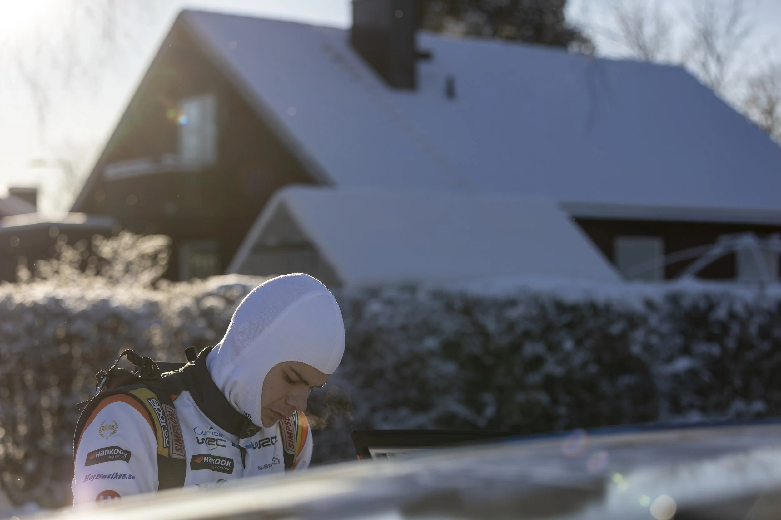 A person dressed in rally racing gear, including a white helmet and a racing suit with various sponsor logos, is outdoors near snow-covered ground with a house and leafless trees in the background, possibly during a winter rally event.