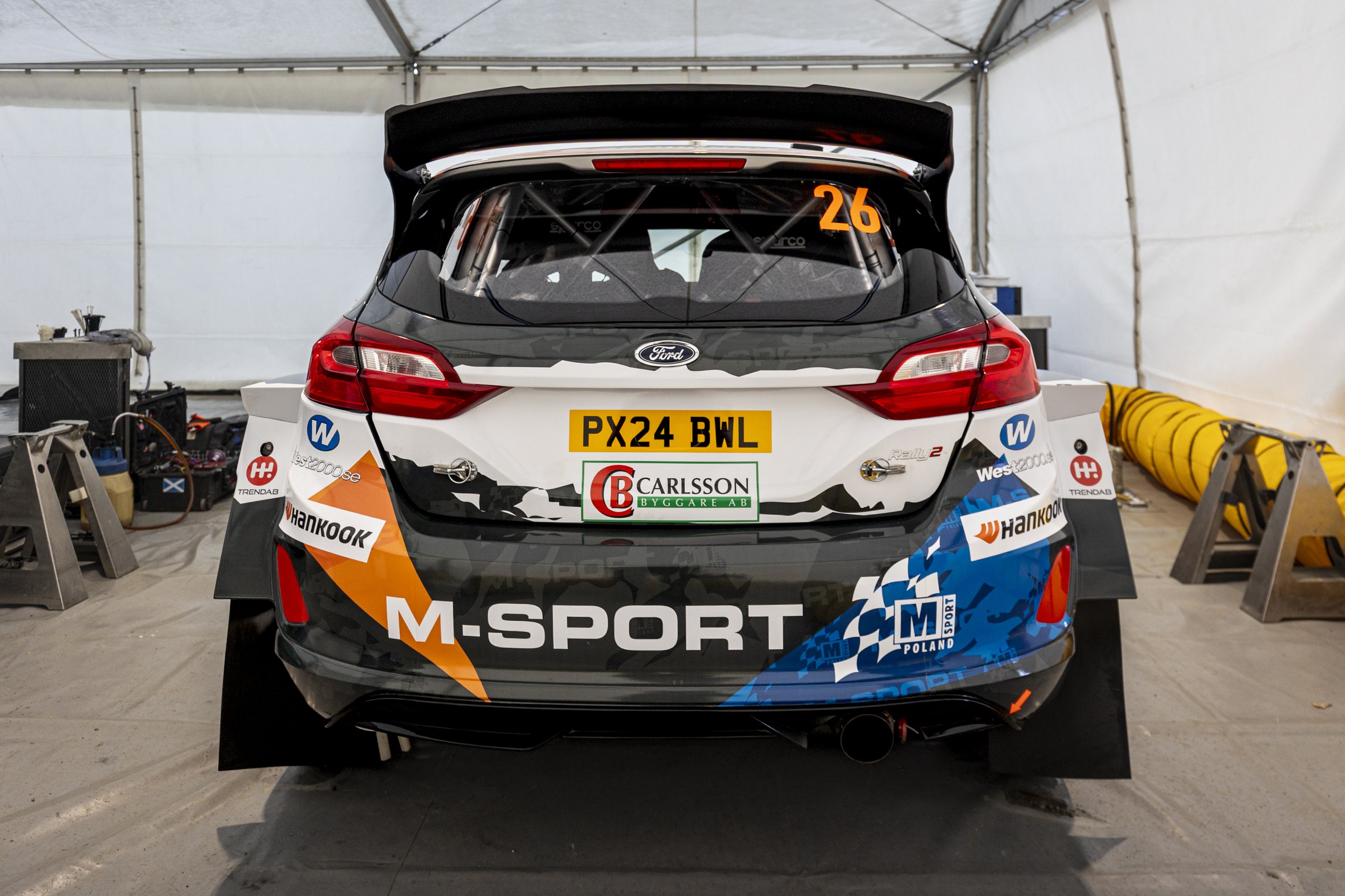 A race car with sponsor logos and decals, inside a tent or garage. The car has the number 26 on the rear window and the license plate PX24 BWL.