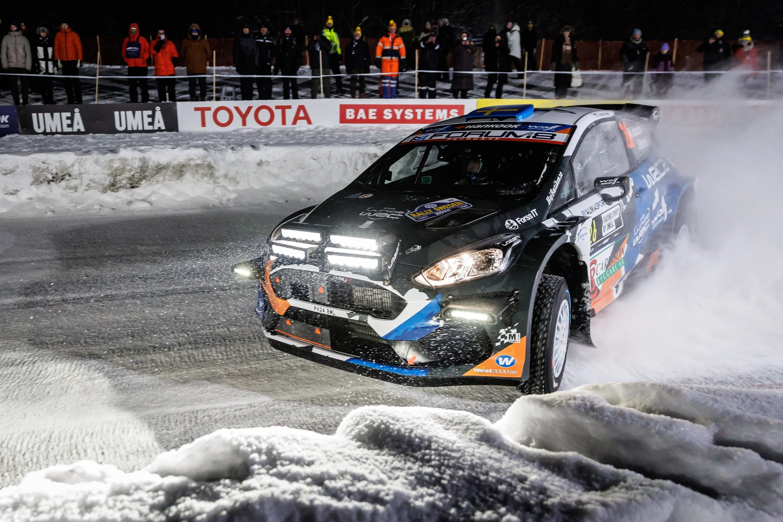 A rally car racing on a snowy track at night, kicking up snow and steam, with spectators watching behind a safety barrier.